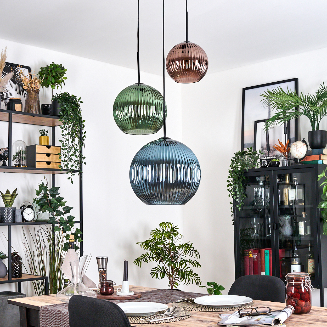 Esszimmer mit drei kugelförmigen Pendelleuchten. Tisch gedeckt mit Tellern, Gläsern und einer Pflanze. Regale und ein Schrank im Hintergrund.