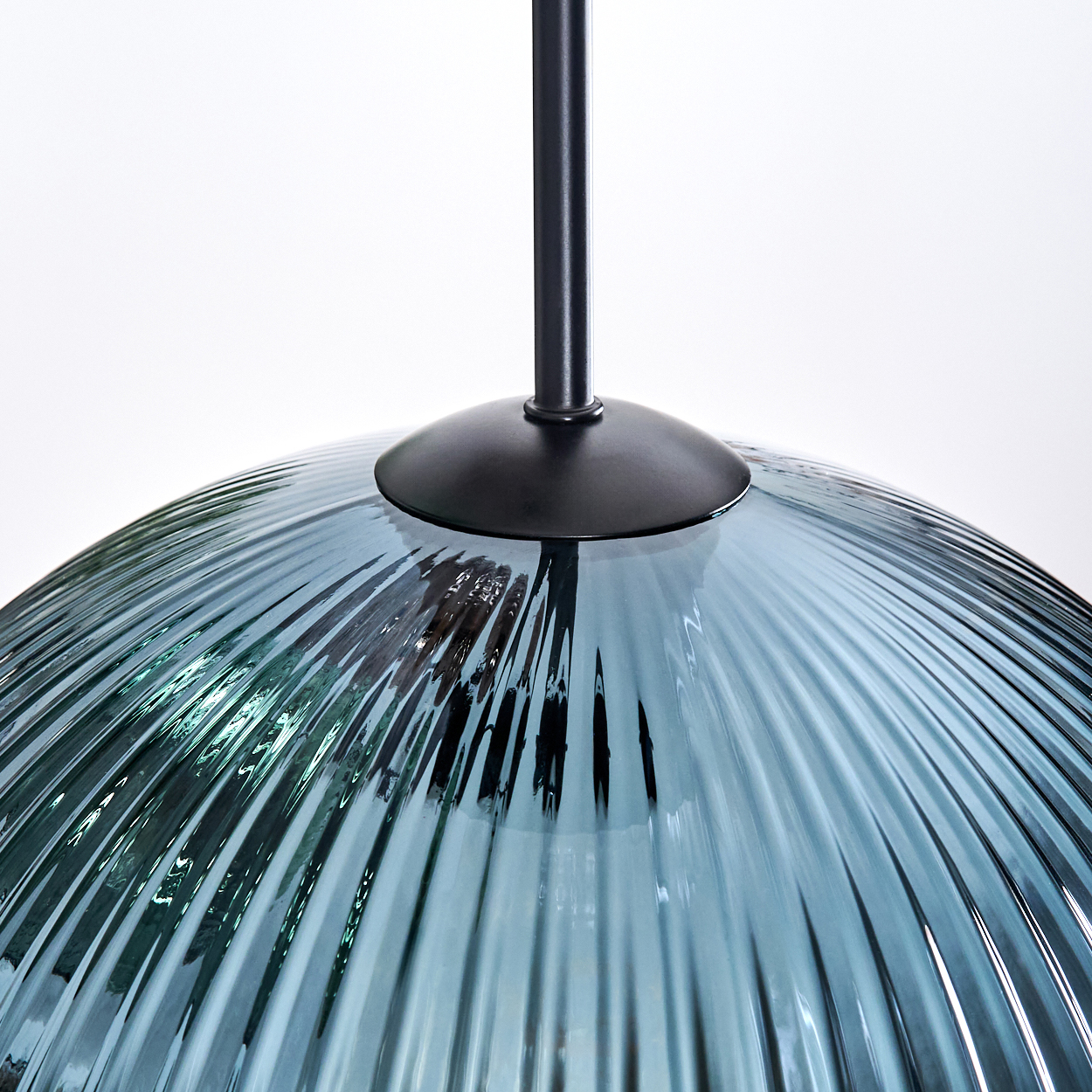 Nahaufnahme einer blauen Glaslampe mit vertikalen Linien. Die Lampe hängt an einer schwarzen Metallstange vor weißem Hintergrund.