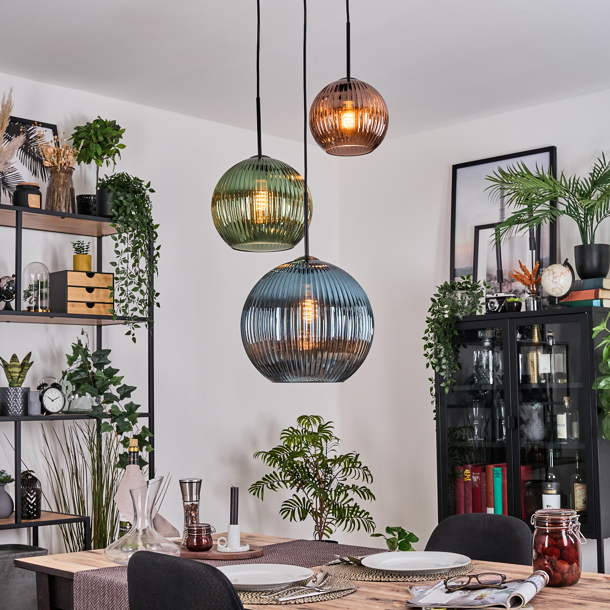 Esszimmer mit Holztisch, drei Hängelampen, Pflanzen und einem schwarzen Schrank mit Dekoration. Teller und Gläser sind gedeckt.
