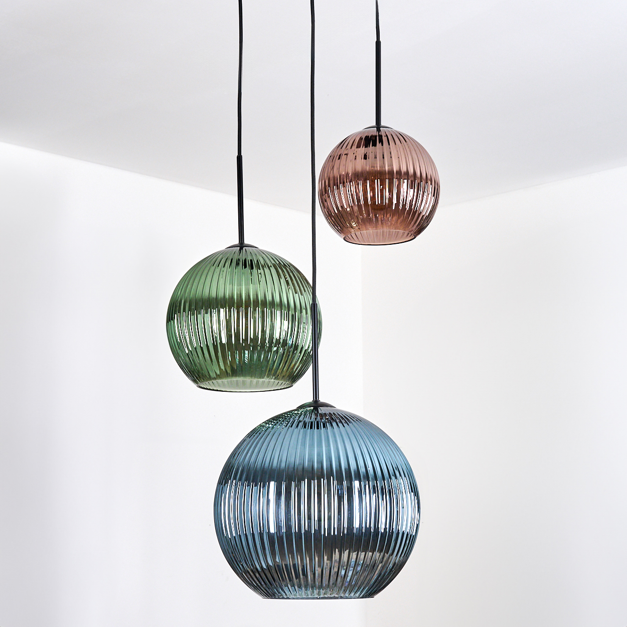 Drei kugelförmige Pendelleuchten in Grün, Rosa und Blau hängen von der Decke. Das gerippte Glas reflektiert Licht, mit weißer Wand im Hintergrund.