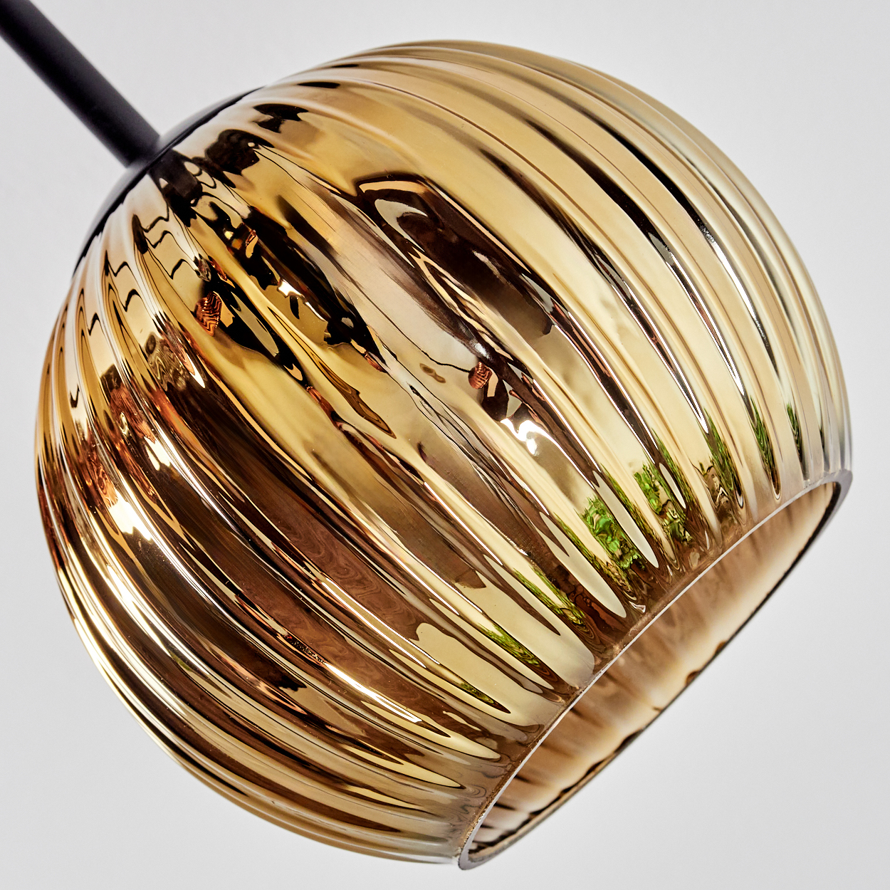 Nahaufnahme eines goldenen, gerippten Lampenschirms aus Glas, der an einem schwarzen Stiel vor weißem Hintergrund hängt.