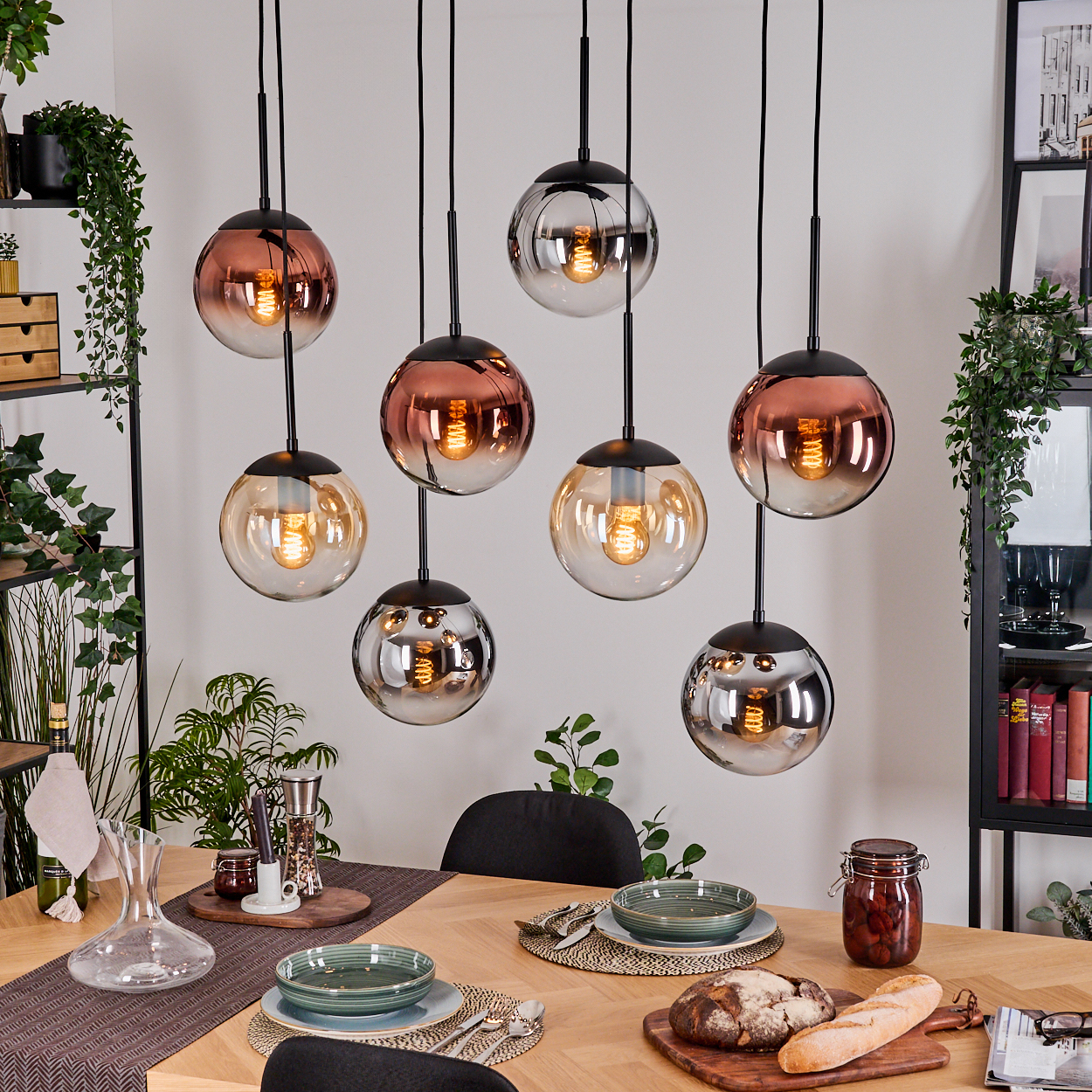 Esszimmer mit hängenden Kugelleuchten. Tisch mit Tellern, Brot und einem Glas mit Essen. Pflanzen und Regale im Hintergrund.