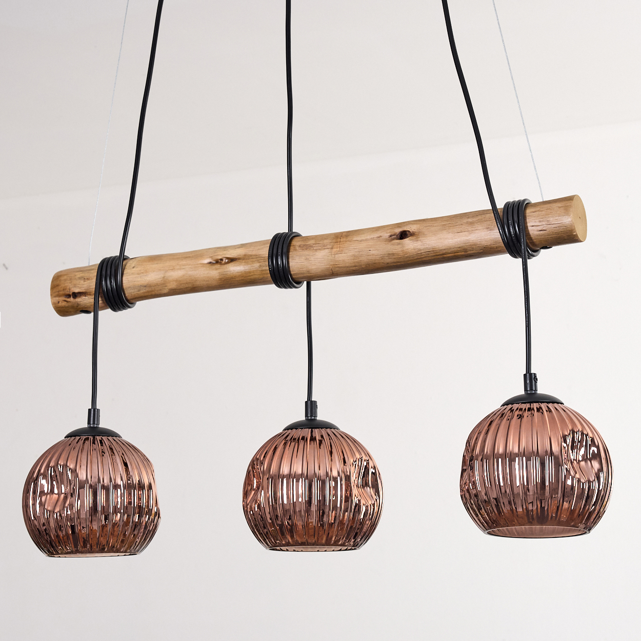 Eine moderne Hängelampe mit drei kupferfarbenen, kugelförmigen Lichtern, die an einem Holzbalken hängen. Schwarze Drähte und weißer Hintergrund.