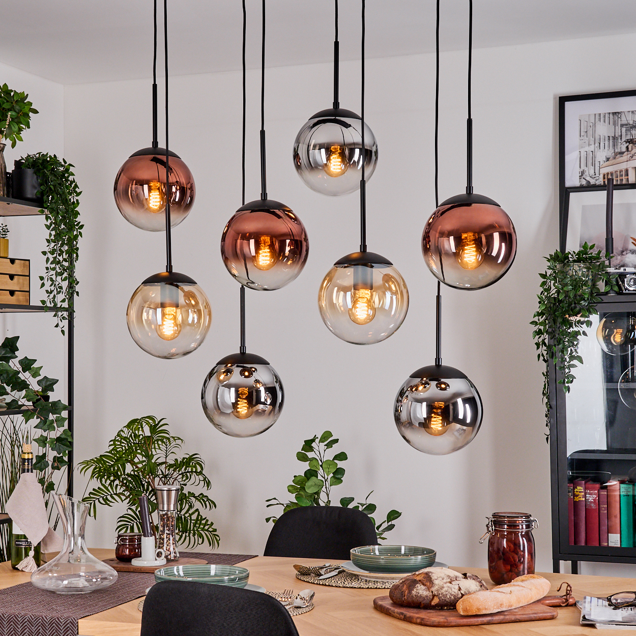 Esszimmer mit mehreren Kugel-Pendelleuchten. Tisch gedeckt mit Essen, Pflanzen und Dekoration. Schwarze Regale mit Büchern und Pflanzen.