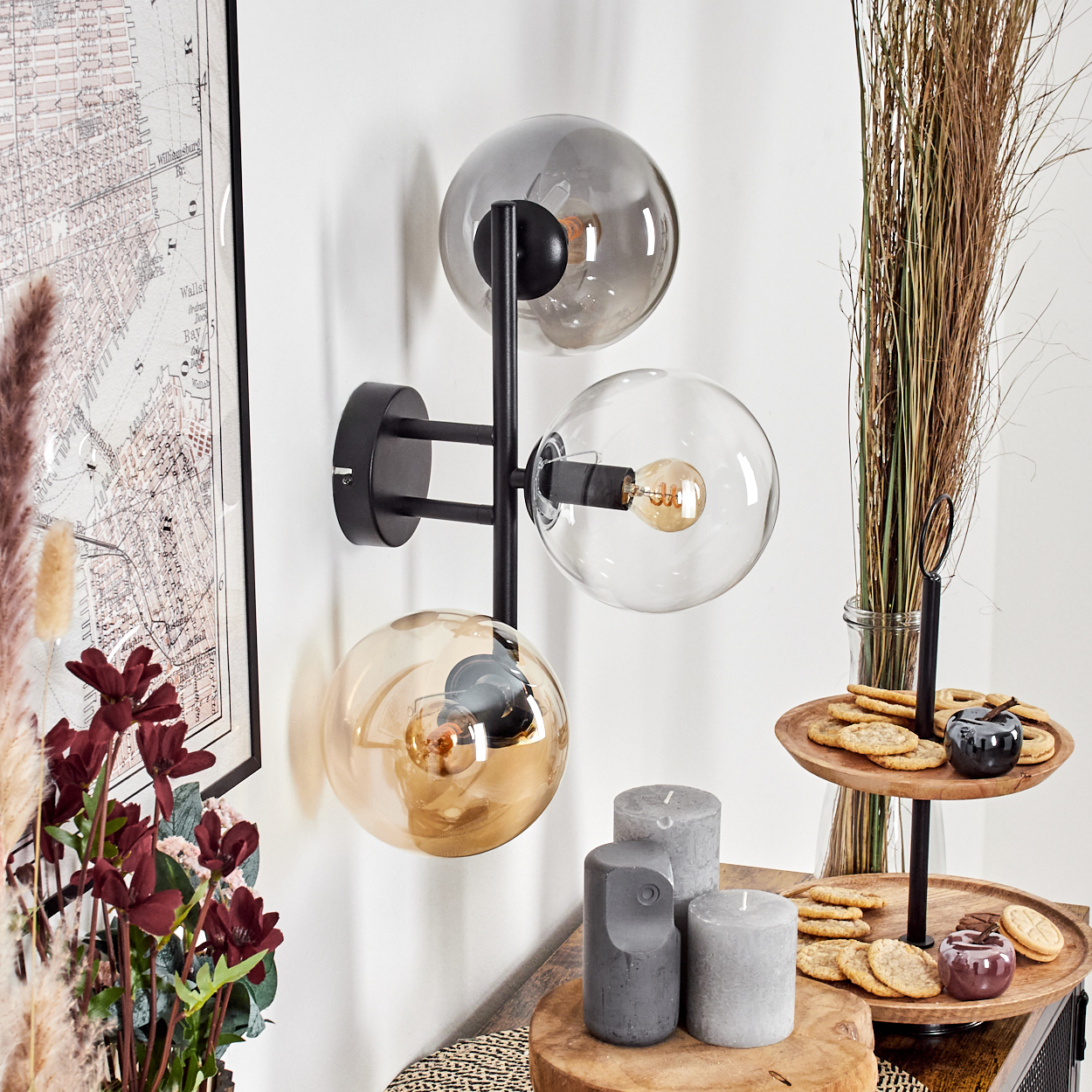 Eine moderne Wandlampe mit drei Glaskugeln. Daneben befinden sich eine Karte, Blumen, Kerzen und ein mehrstöckiges Tablett mit Keksen.