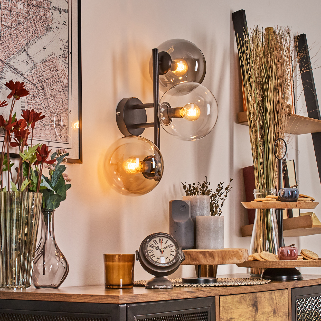 Ein Zimmer mit Wandlampe, Uhr, Blumen und Dekorationsgegenständen auf einem Holzschrank. An der Wand ist eine Karte zu sehen.