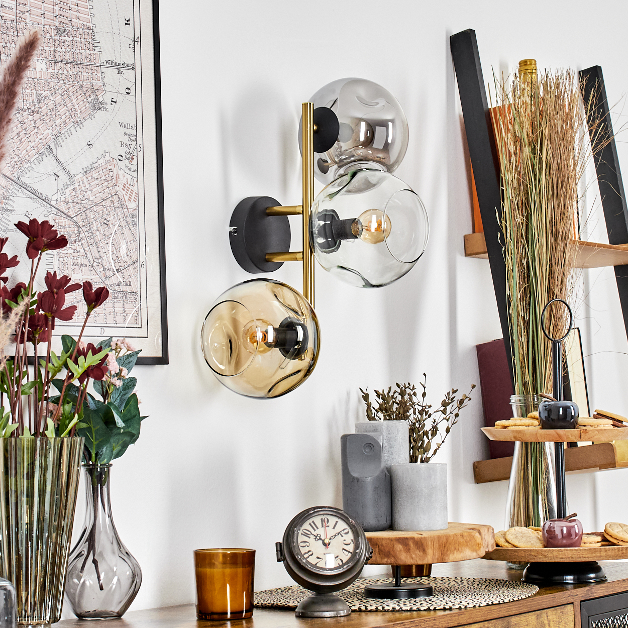 Eine Wandlampe mit drei Glaskugeln. Eine Karte, Blumen, eine Uhr und dekorative Gegenstände stehen auf einer Holzoberfläche.