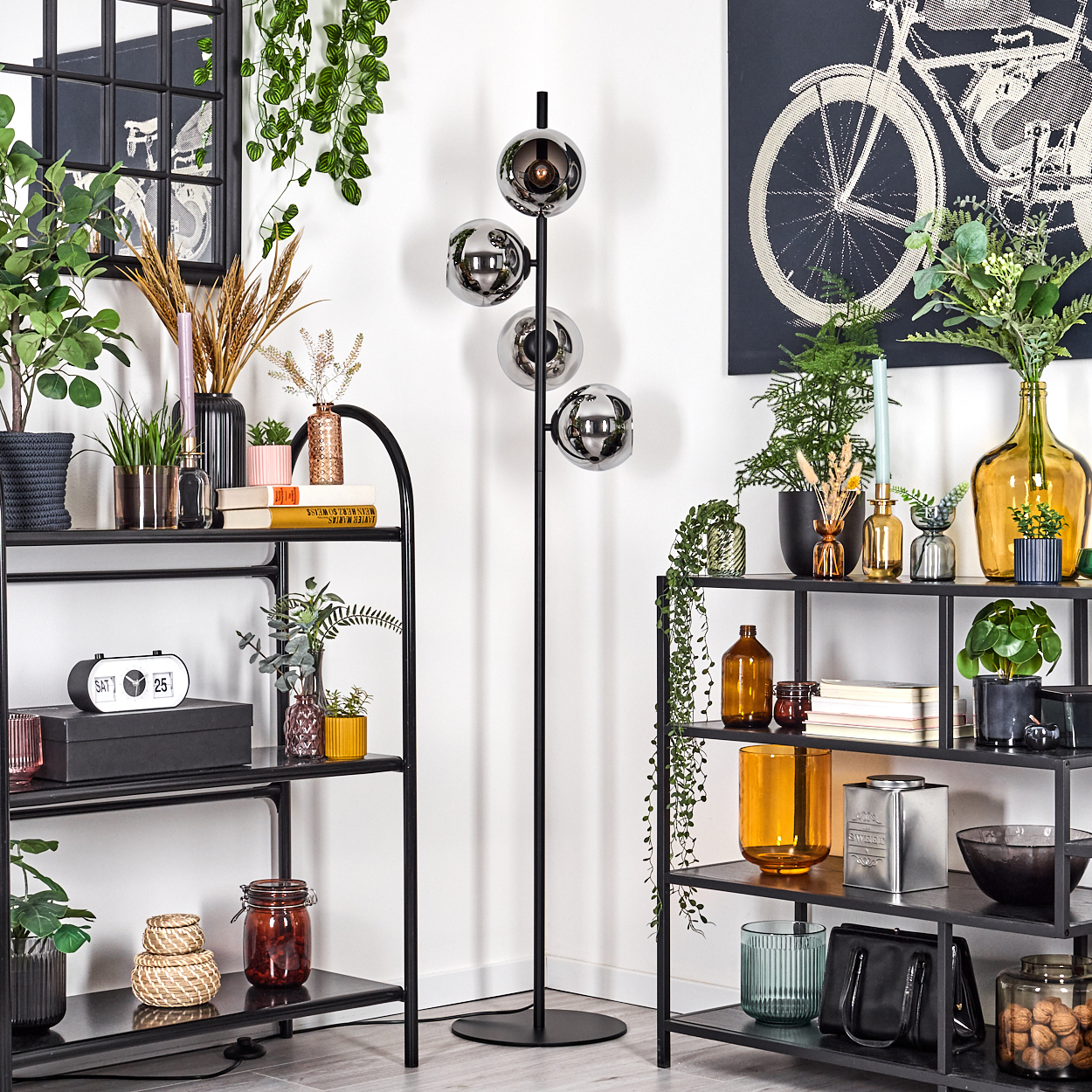 Ein Raum mit Regalen, Pflanzen und einer schwarzen Stehlampe mit Glaskugeln. Ein Fahrrad-Druck hängt an der Wand.