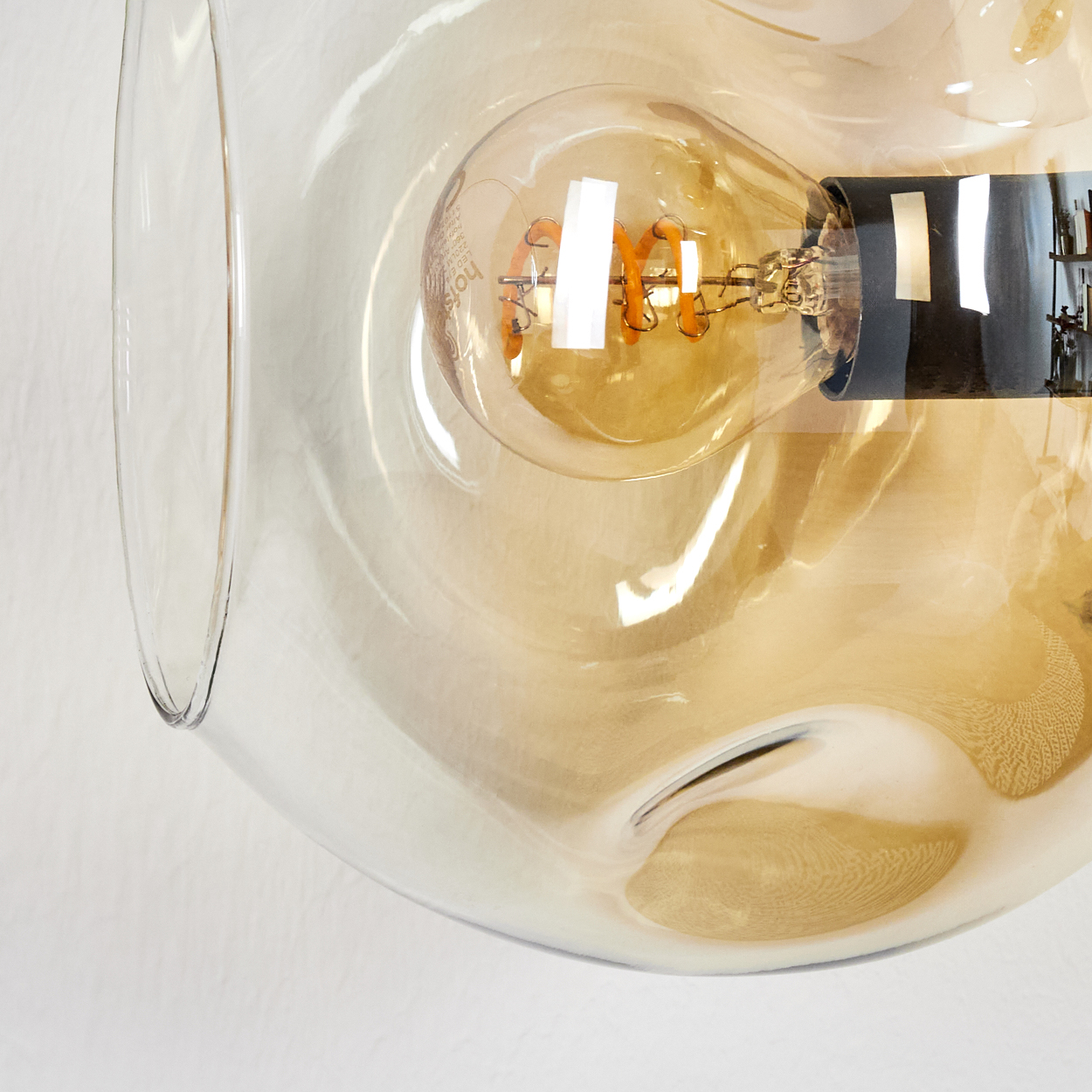 Nahaufnahme einer Glasleuchte mit sichtbarer Glühbirne. Das Glas ist klar mit Goldtönung, an einer weißen Wand befestigt.
