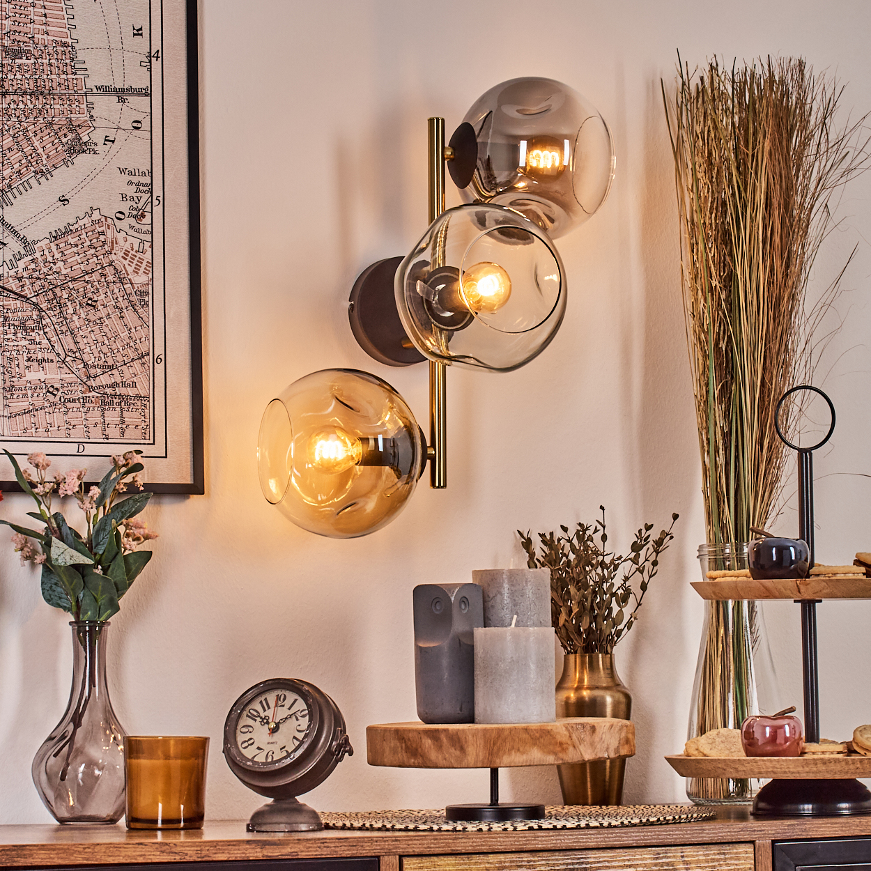 Eine Wandlampe mit drei Glaskugeln. Verschiedene Dekorationsgegenstände, darunter Kerzen, Uhr, Vase und Karte, sind auf einer Holzoberfläche angeordnet.