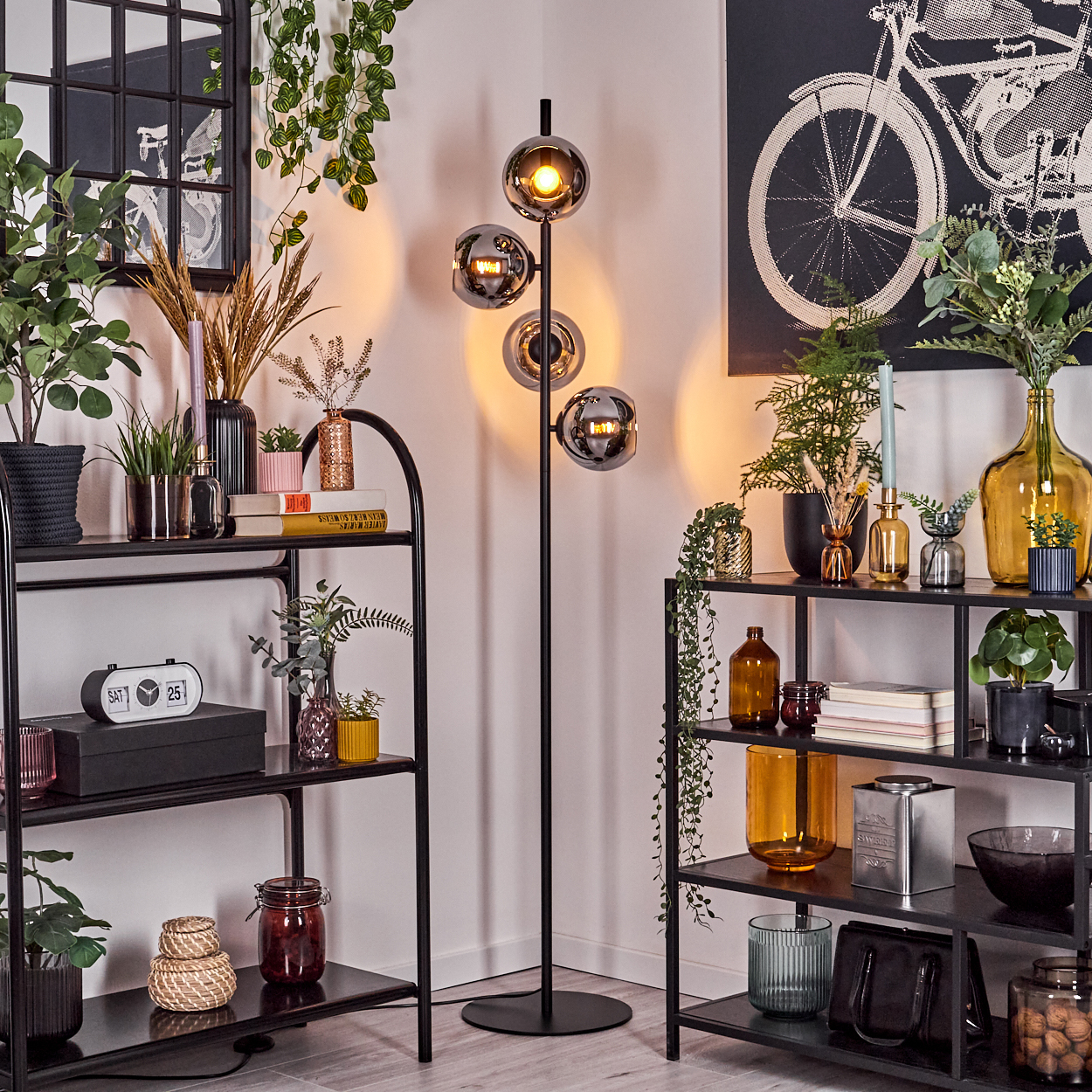 Raumecke mit Regalen, Pflanzen und einer hohen Lampe. Ein Fahrrad-Kunstdruck hängt an der Wand und sorgt für Dekoration.