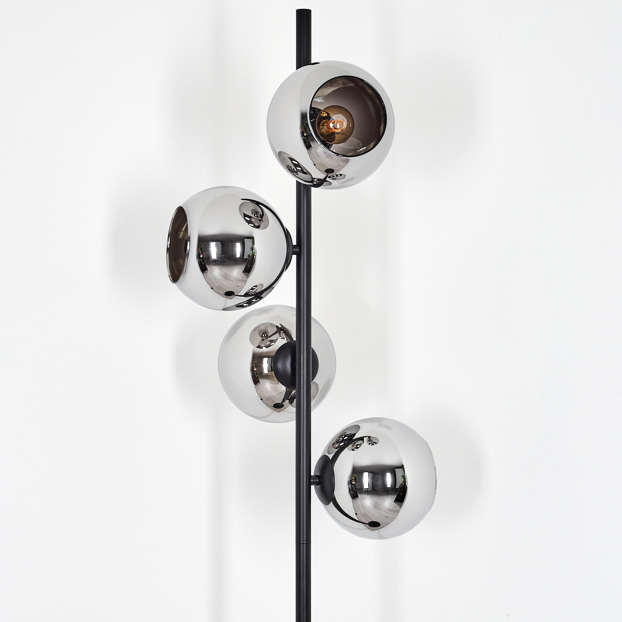 Eine moderne Stehlampe mit vier verchromten Kugelschirmen. Ein schwarzer Pfosten trägt die Kugeln vor einer weißen Wand.