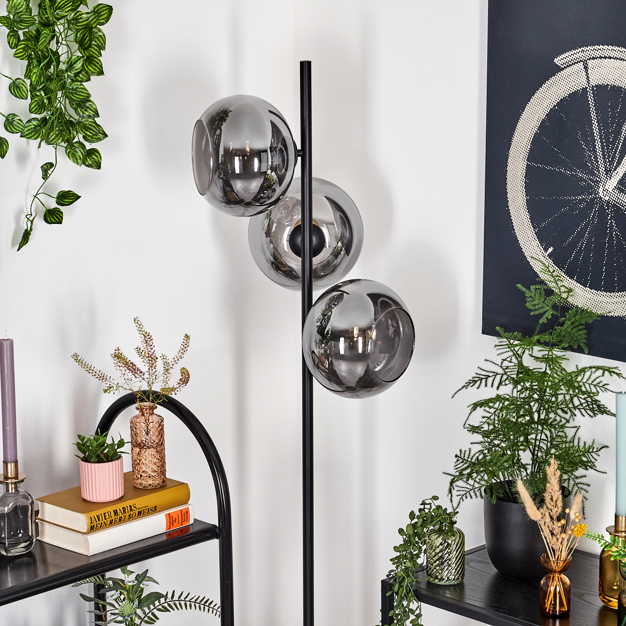 Ein modernes Zimmer mit einer schwarzen Lampe mit drei Glaskugeln. Es gibt Pflanzen, Regale und ein Fahrradrad-Kunstwerk.