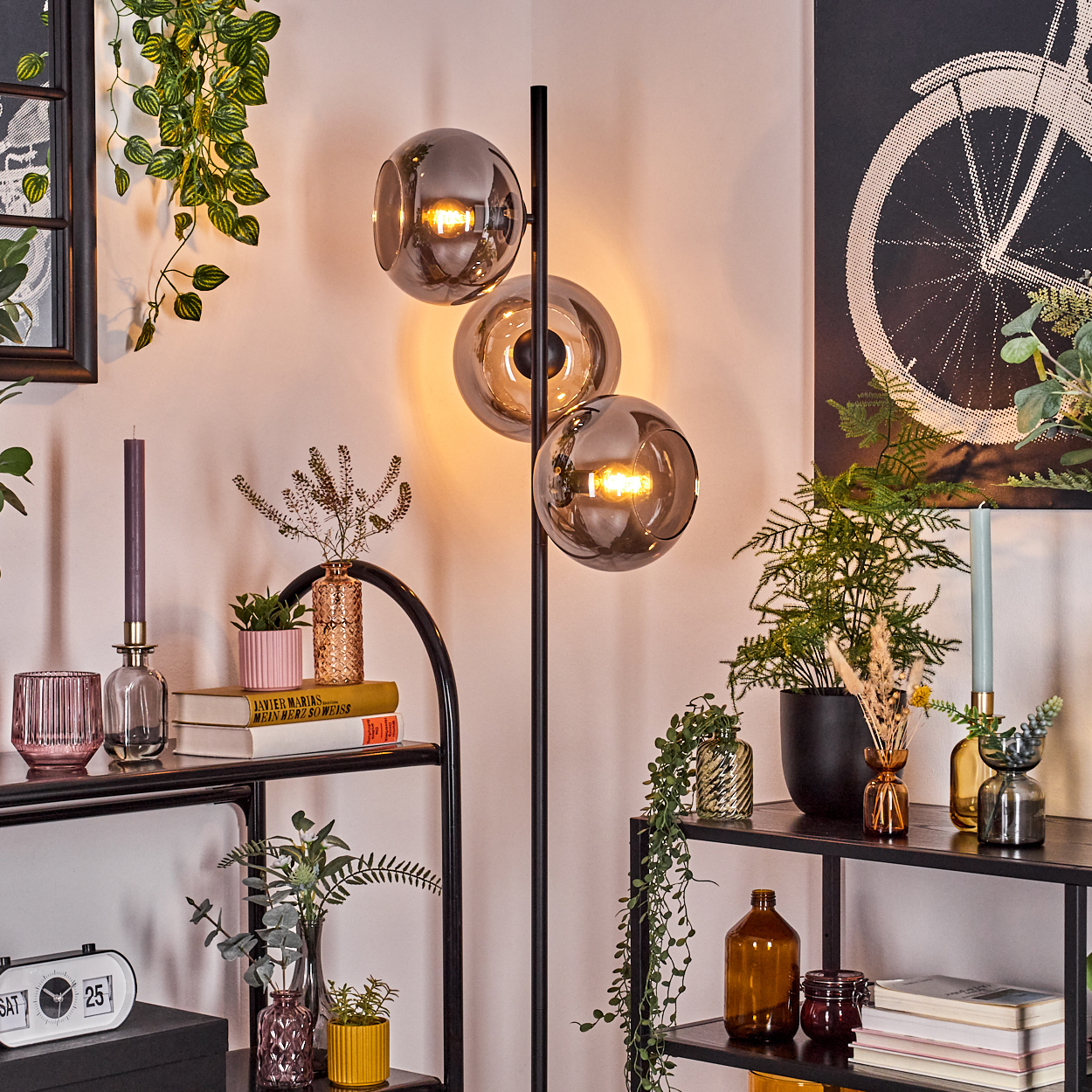 Moderne Wohnzimmerecke. Eine hohe Lampe mit drei Glaskugeln. Regale zeigen Pflanzen, Bücher und Kerzen. Ein Fahrradkunstwerk hängt an der Wand.
