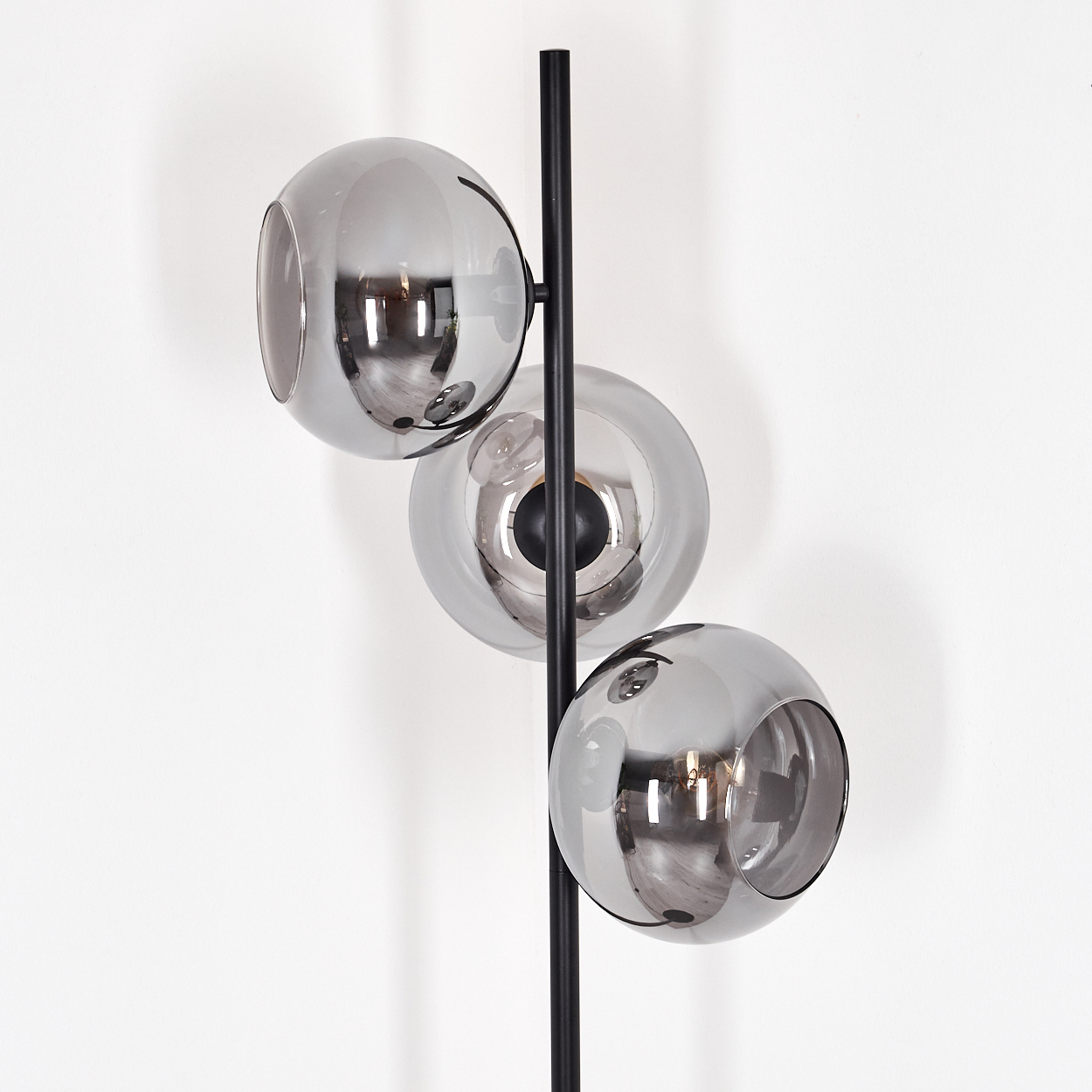 Eine moderne Lampe mit drei grauen Glaskugelschirmchen an einem schwarzen Mast vor einer weißen Wand. Licht spiegelt sich im Glas.
