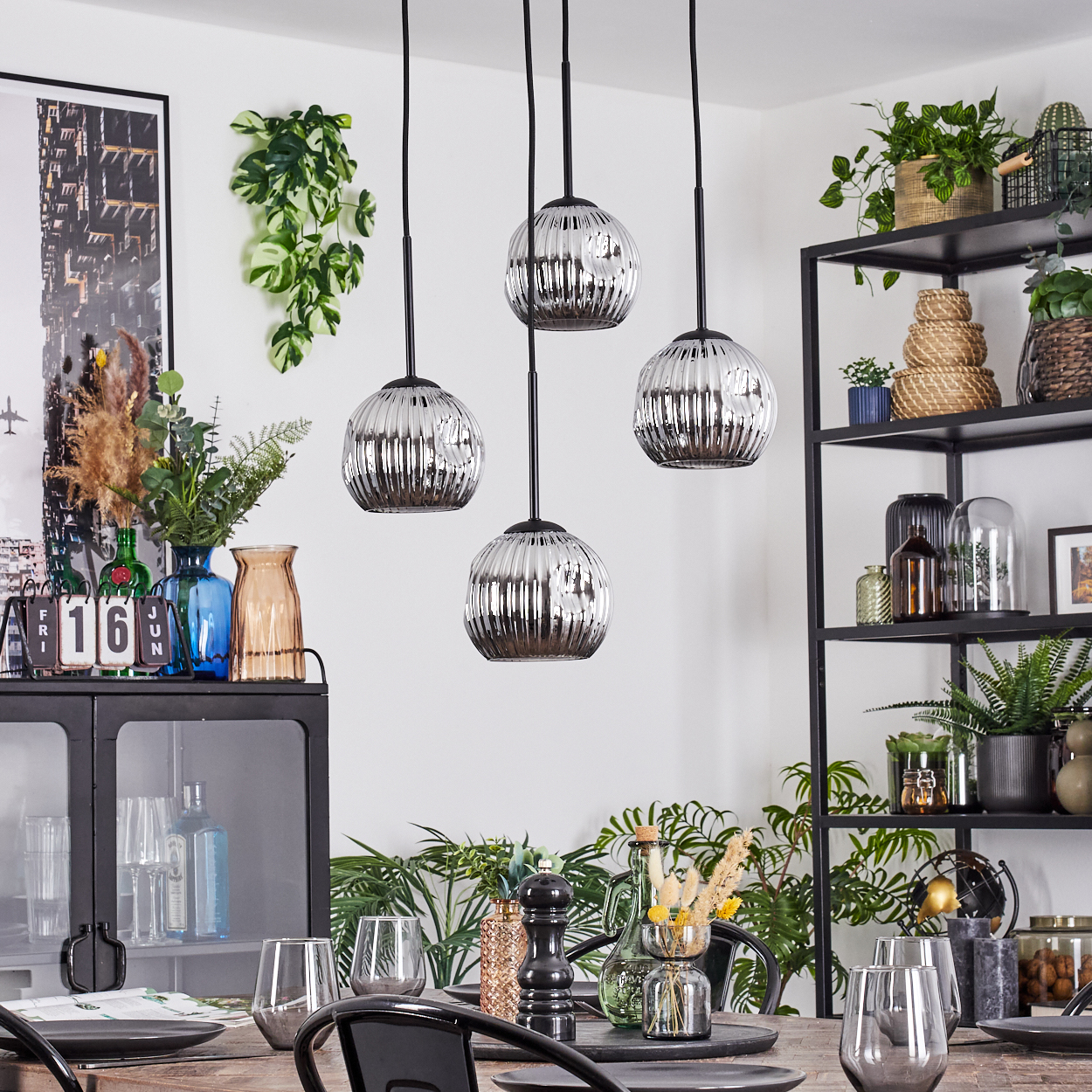 Esszimmer mit Hängelampen, Pflanzen und Regalen. Tisch mit Tellern und Gläsern. Schrank und Dekorationsartikel sichtbar.