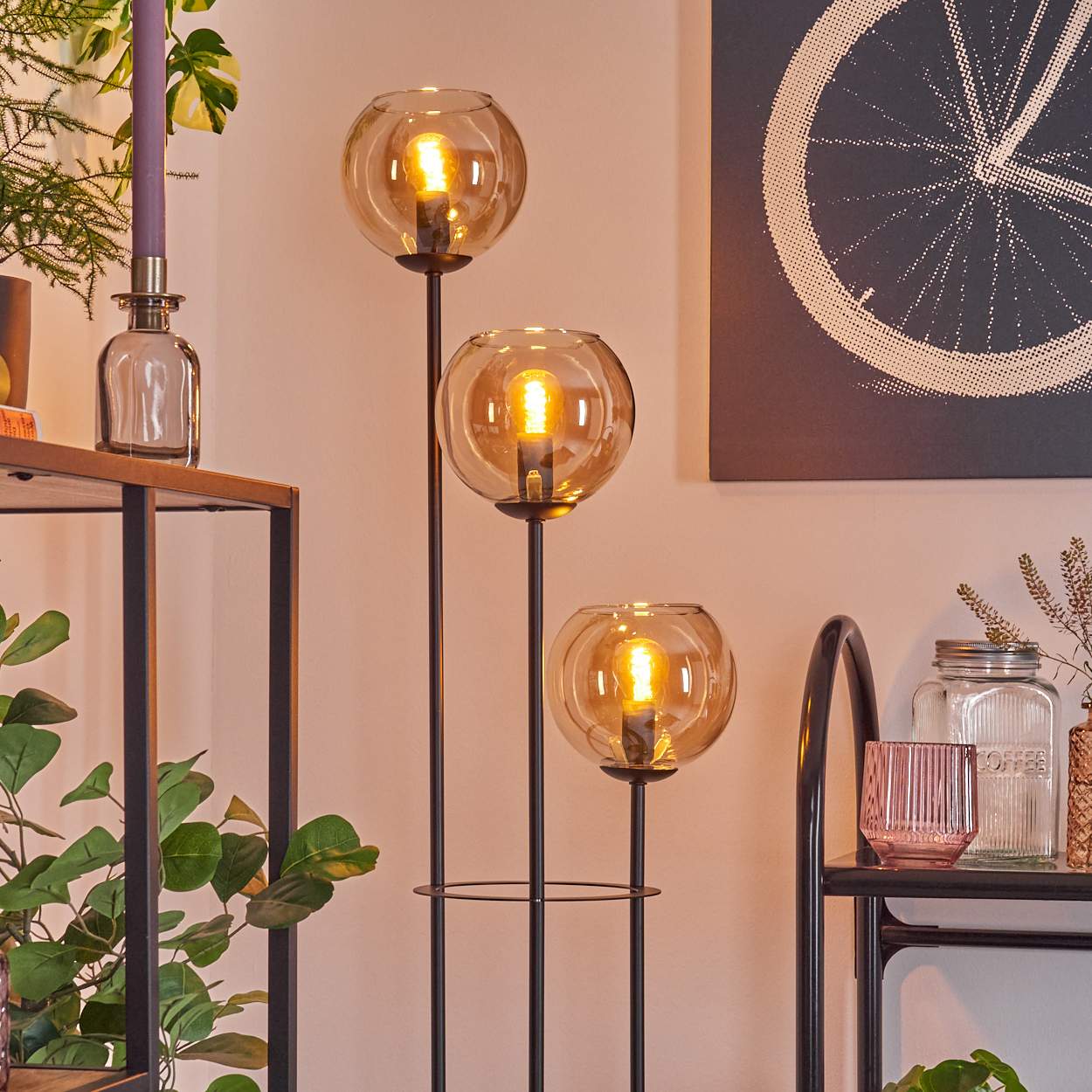 Eine Stehlampe mit drei Glaskugelschirmen. Im Hintergrund befinden sich eine Kerze, Pflanzen und ein Fahrradrad-Kunstwerk.