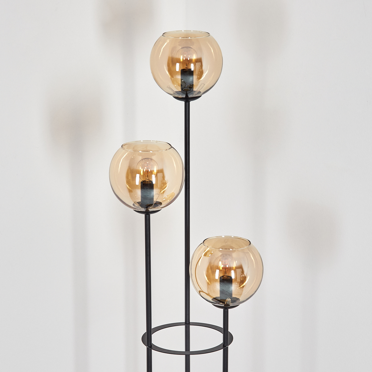 Eine moderne Stehlampe mit drei kugelförmigen, bernsteinfarbenen Glasschirmen, jeweils mit Glühbirne, auf schwarzen Metallstielen an einer weißen Wand.