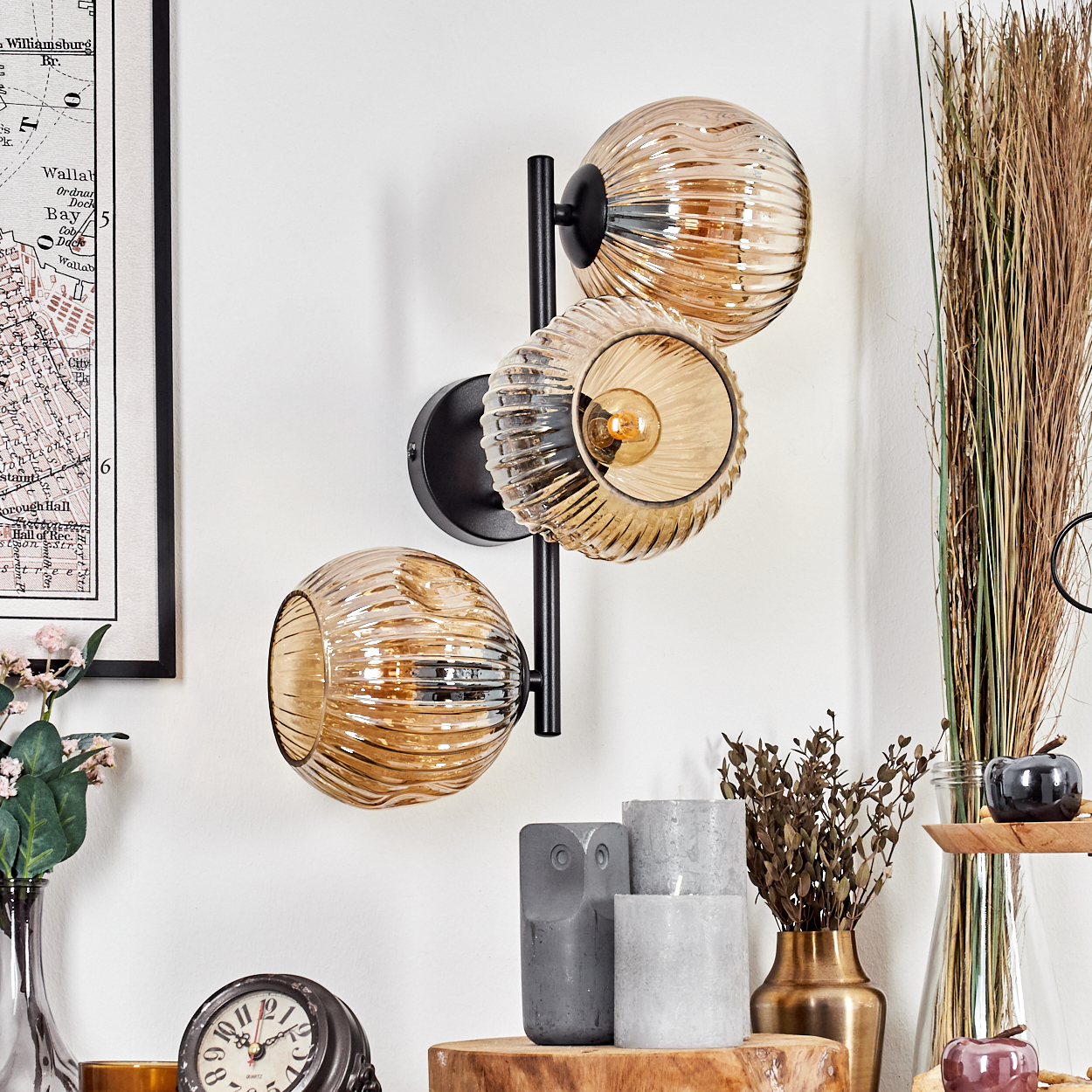 Eine Wandlampe mit drei bernsteinfarbenen Glaskugeln an einer weißen Wand. Darunter Kerzen, Uhr und Vase mit getrockneten Pflanzen.