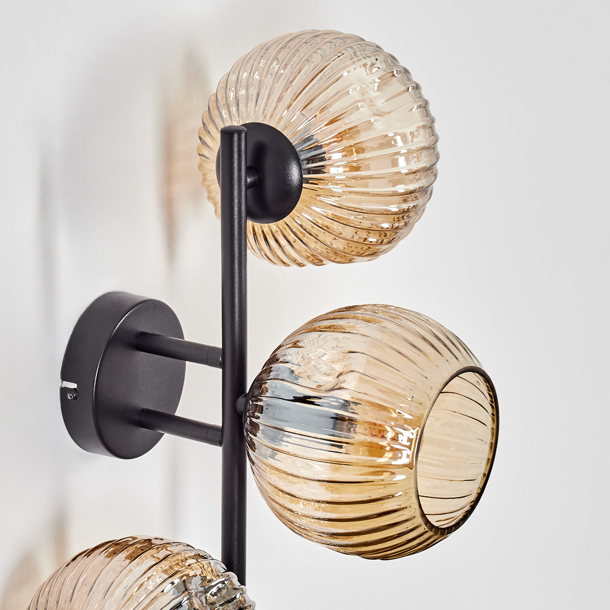 Eine moderne Wandlampe mit drei kugelförmigen, bernsteinfarbenen Glasschirmen. Die Lampe hat ein schwarzes Metallgestell an einer weißen Wand.
