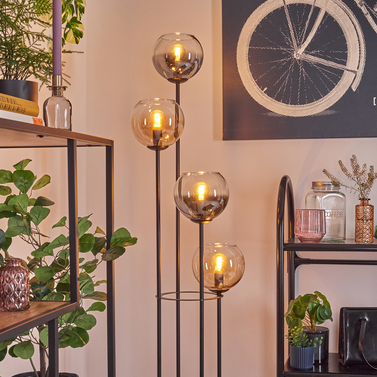 Eine moderne Stehlampe mit vier Glaskugeln, beleuchtet, steht in einem Raum mit Regalen und Pflanzen. Ein Fahrradrad-Kunstwerk an der Wand.