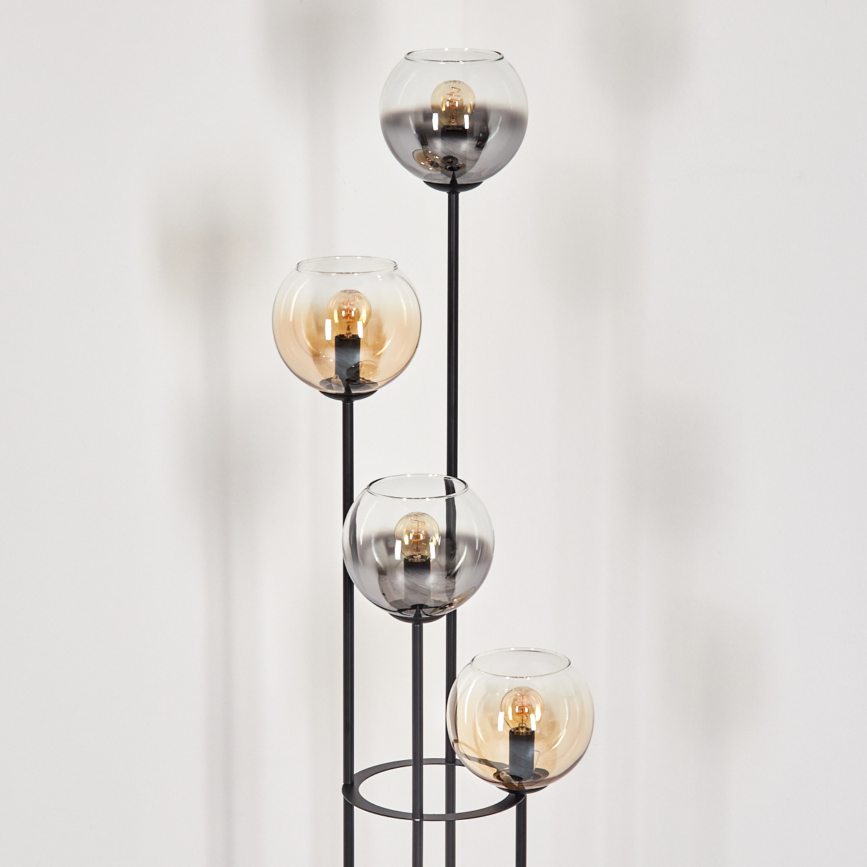 Eine moderne Stehlampe mit vier Glaskugel-Schirmen, in denen sich jeweils eine Glühbirne befindet. Der Rahmen ist schwarz, der Hintergrund ist eine weiße Wand.