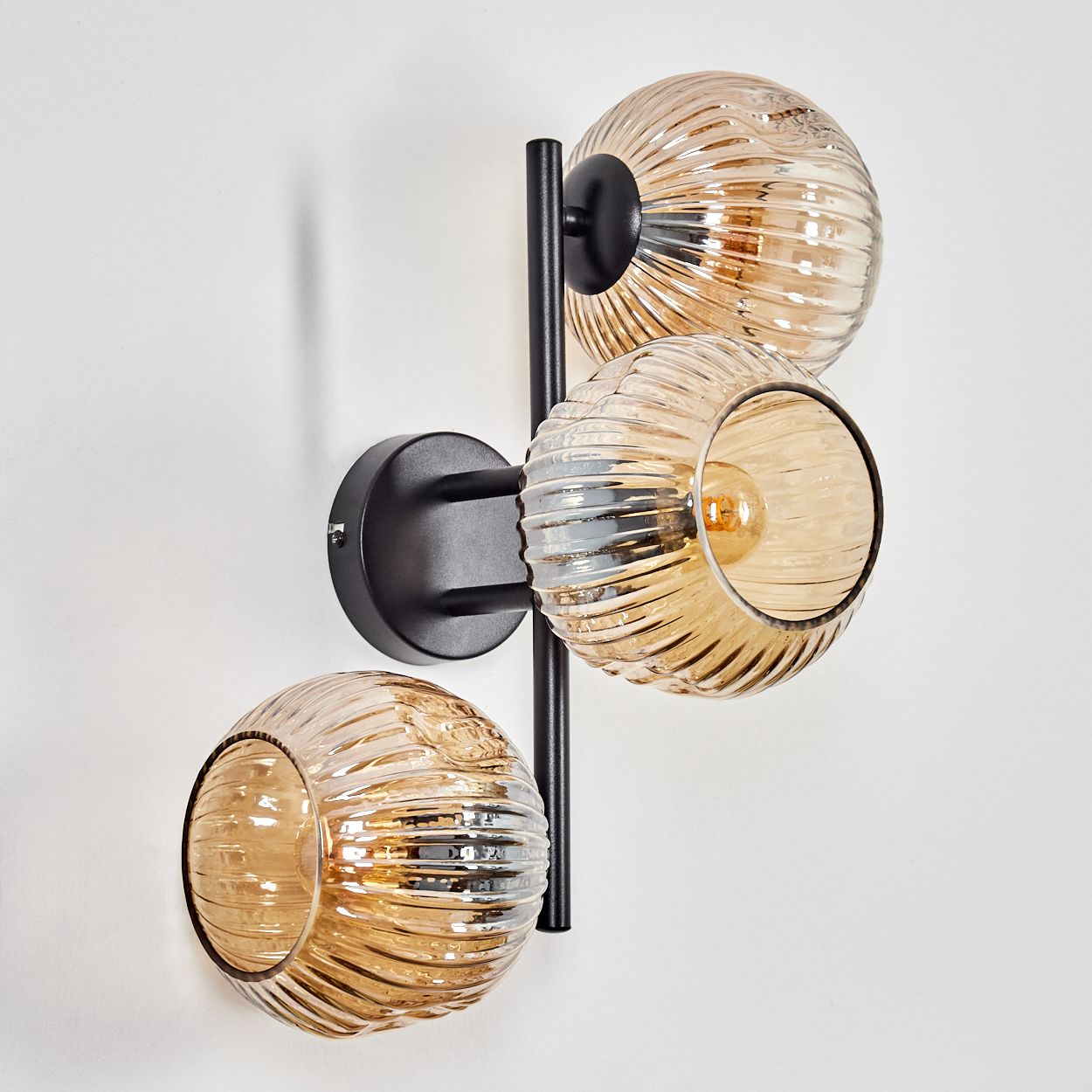 Eine Wandlampe mit drei Glaskugeln. Die Kugeln sind bernsteinfarben und gerippt. Die Halterung ist schwarz und an einer weißen Wand befestigt.