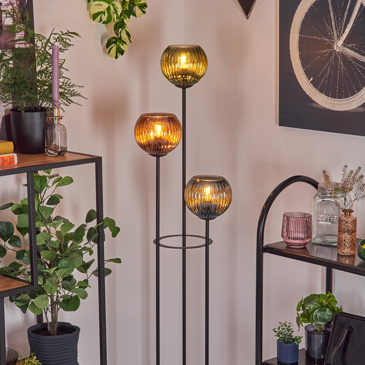 Eine Stehlampe mit drei Glühbirnen und Glasschirmen steht in der Nähe von Regalen mit Pflanzen und Dekoration. Ein Fahrradrad-Kunstwerk hängt an der Wand.