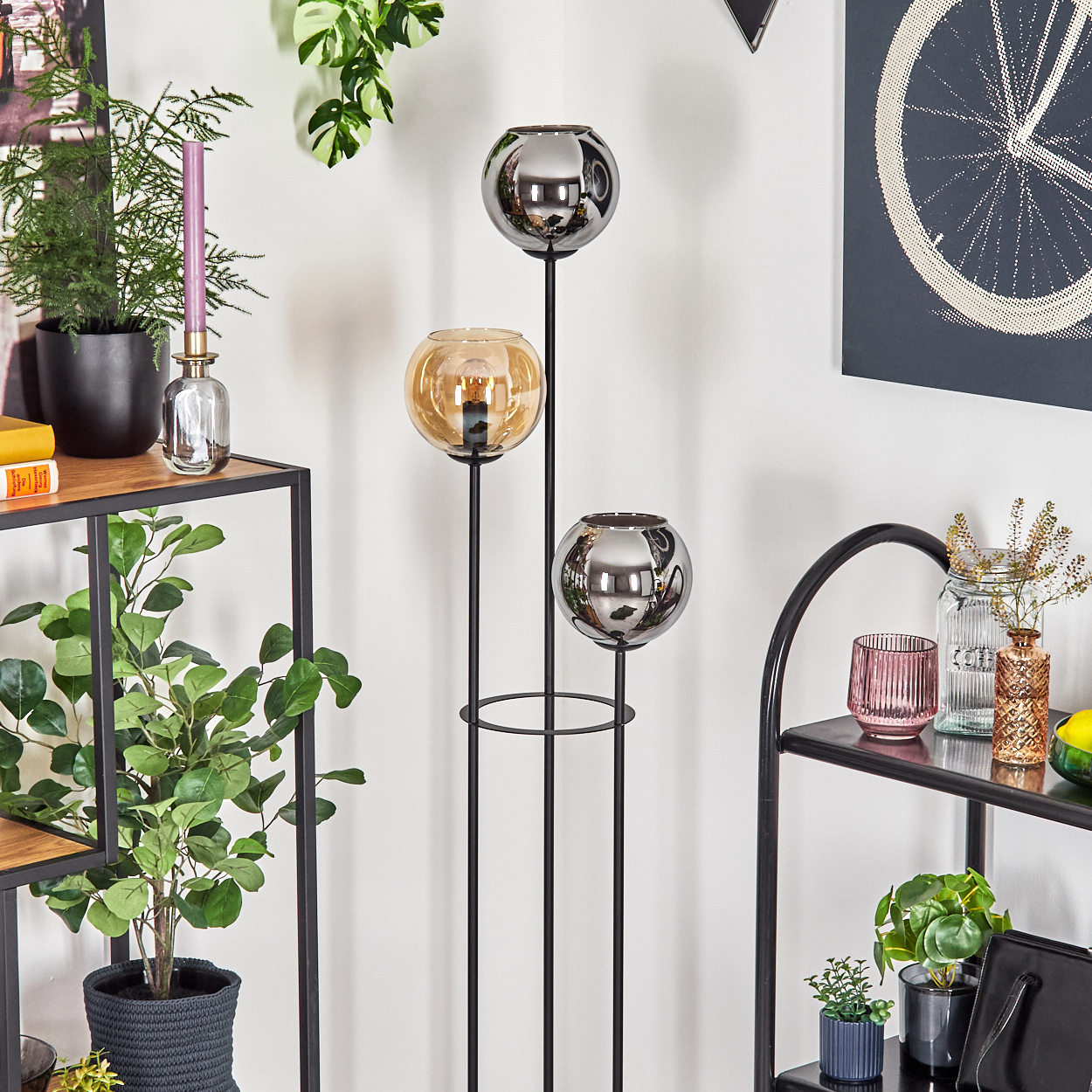 Lampe mit drei Kugeln, Pflanzen, Regale und Kunstwerk an einer weißen Wand. Die Lampe hat silberne und bernsteinfarbene Kugeln.