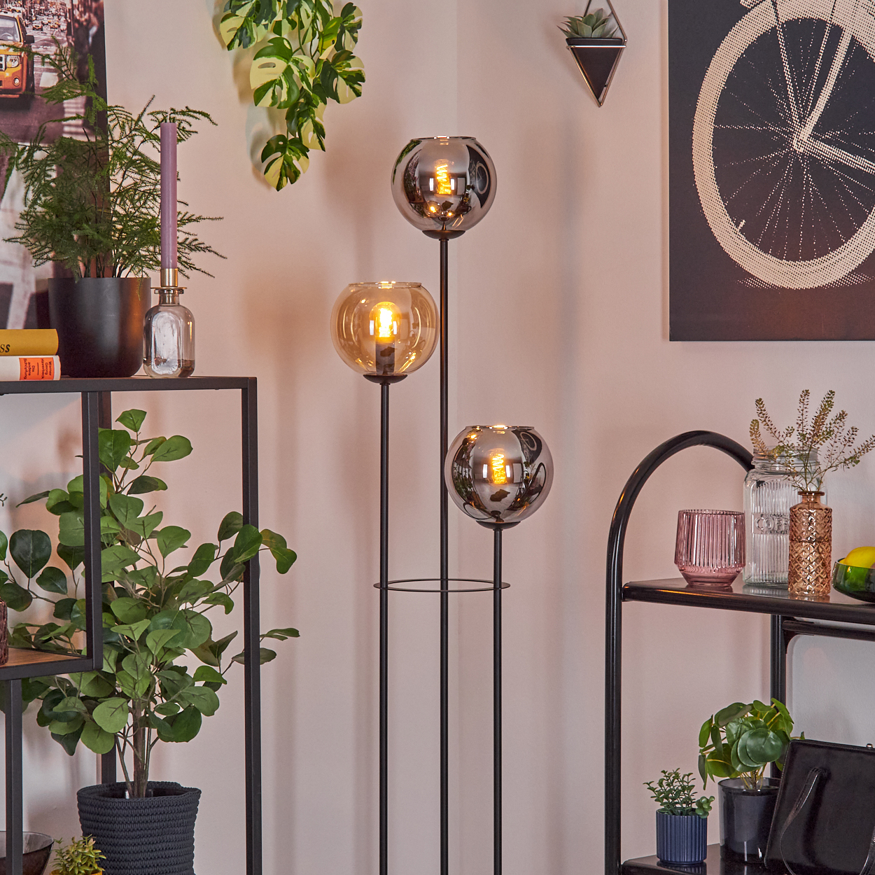 Eine Zimmerecke mit einer schwarzen Metalllampe mit drei Glaskugeln. Regale mit Pflanzen und Dekoration vor hellrosa Wand.