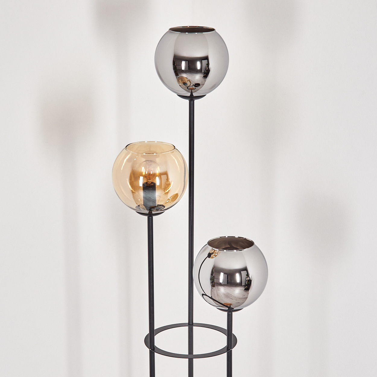 Moderne Lampe mit drei kugelförmigen Schirmen. Zwei sind silber, einer bernsteinfarben. Schwarzer Metallrahmen. Weißer Hintergrund.