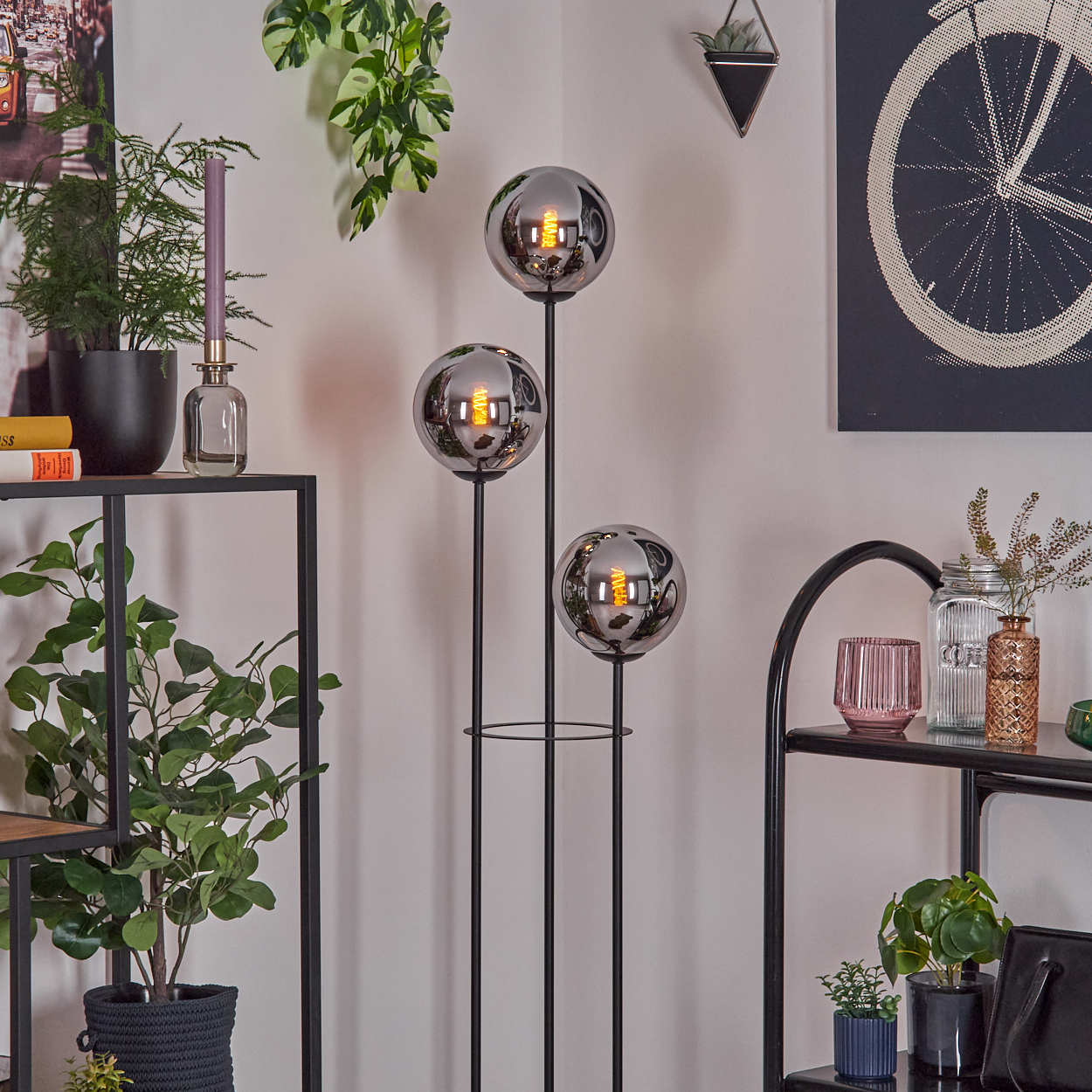 Ein modernes Interieur mit einer schwarzen Metall-Stehlampe mit drei kugelförmigen Lichtern. Pflanzen und dekorative Gegenstände sind ebenfalls zu sehen.