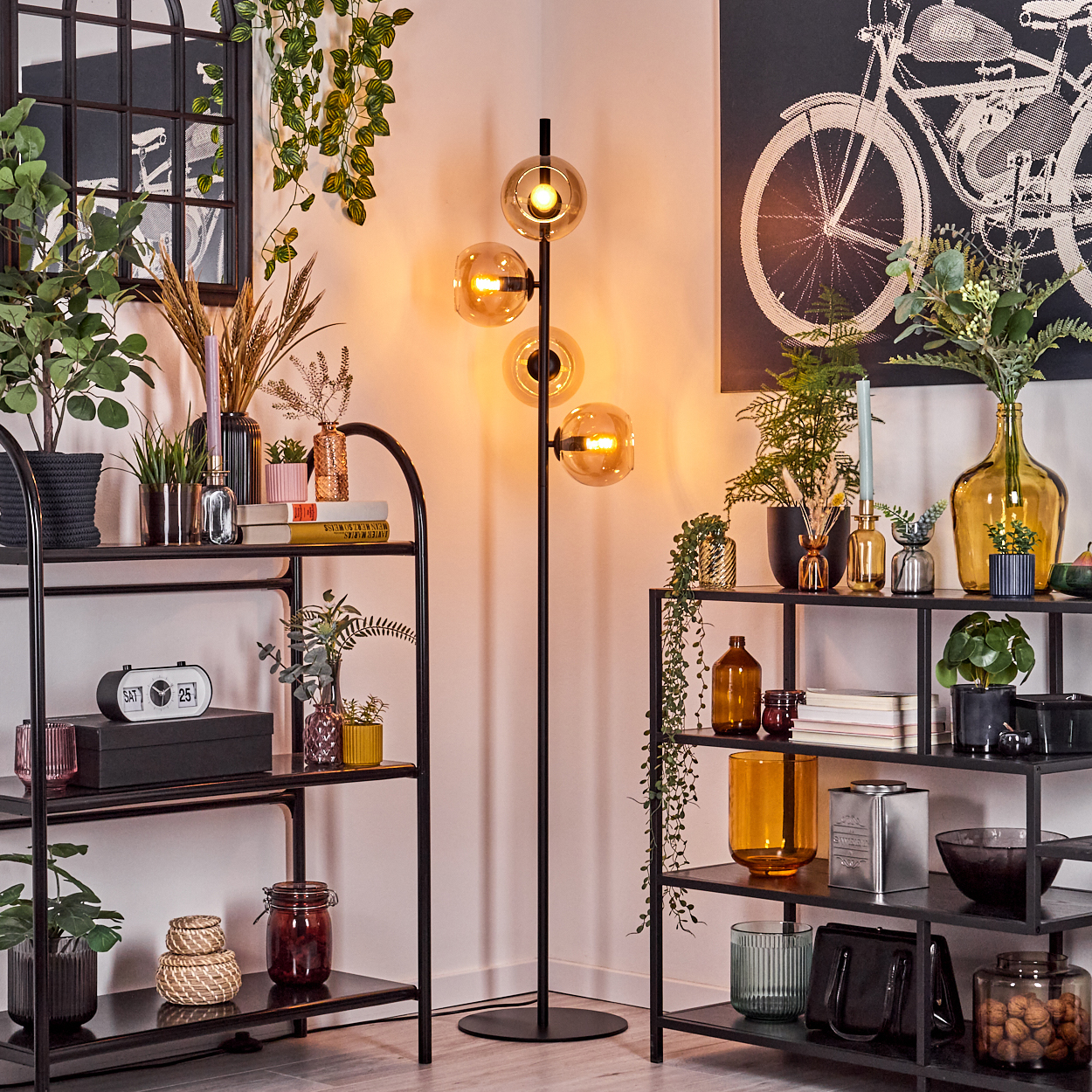 Ein Raum mit Regalen, Pflanzen und einer Stehlampe mit bernsteinfarbenen Glaskugeln. Ein Kunstdruck eines Fahrrads hängt an der Wand.