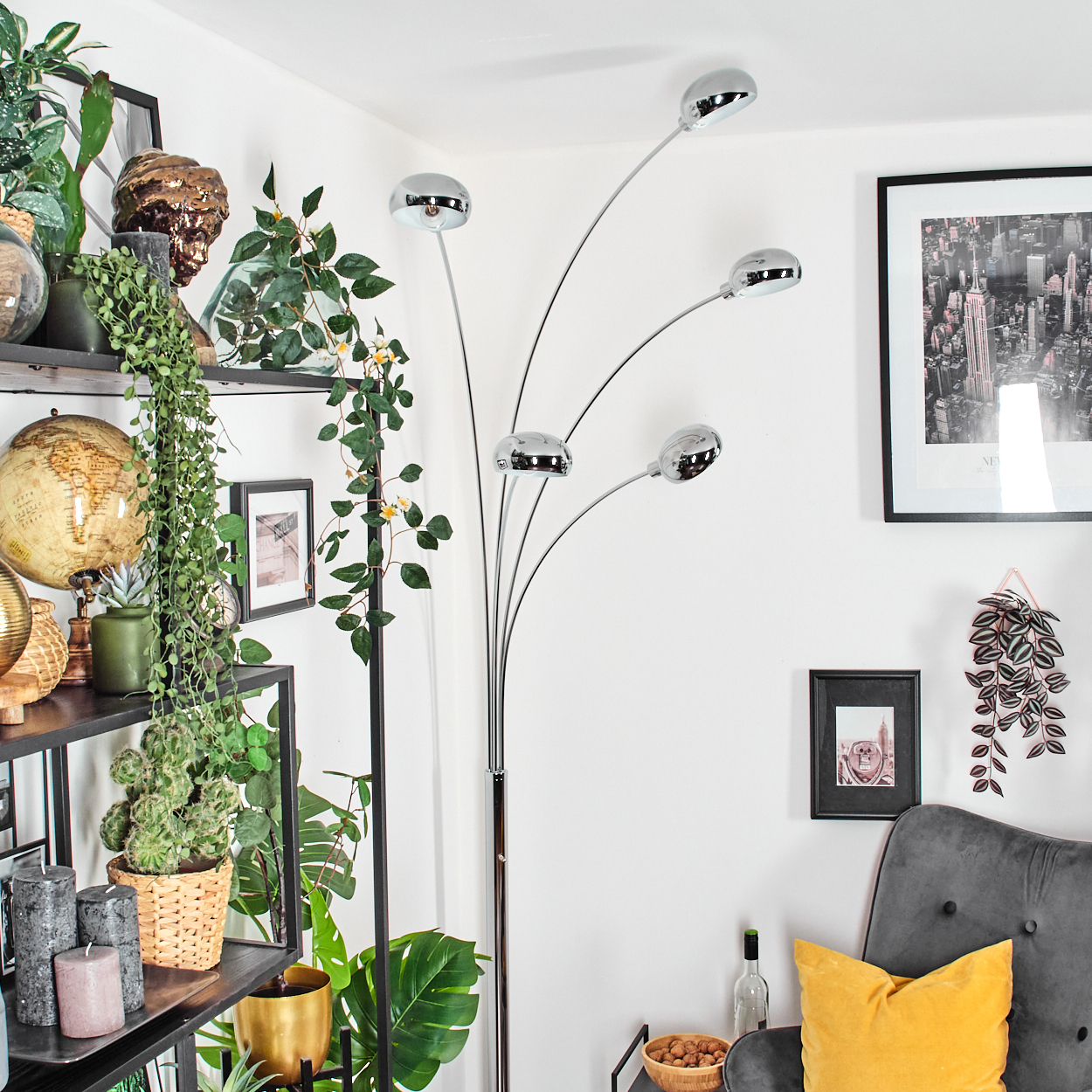 Ein modernes Wohnzimmer mit einer Chrom-Stehlampe, Regalen mit Pflanzen, gerahmten Bildern und einem grauen Sessel mit gelbem Kissen.