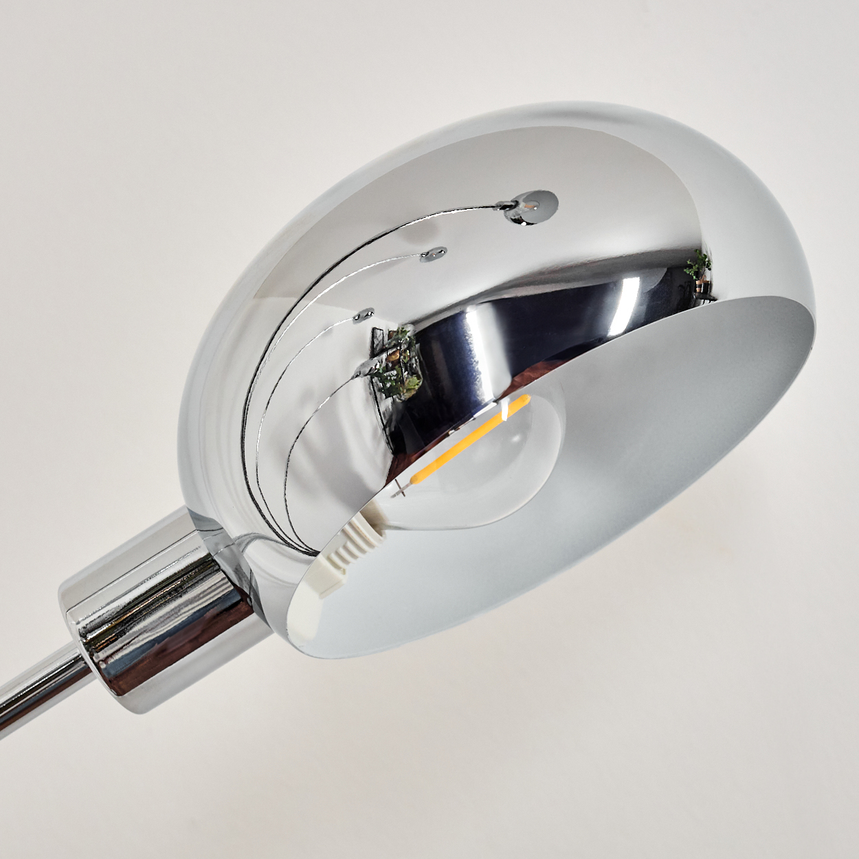 Nahaufnahme eines glänzenden Chrom-Lampenkopfes. Die Lampe reflektiert die Umgebung. Eine freiliegende Glühbirne ist sichtbar.