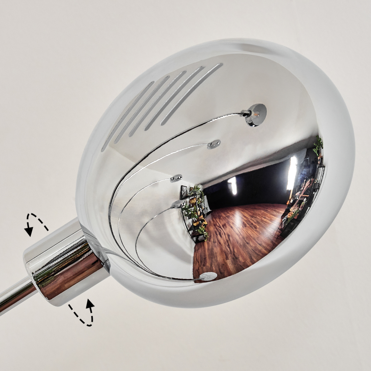 Ein verchromter Lampenkopf spiegelt einen Raum mit Holzböden, Pflanzen und Kunst wider. Die Lampe befindet sich auf weißem Hintergrund.
