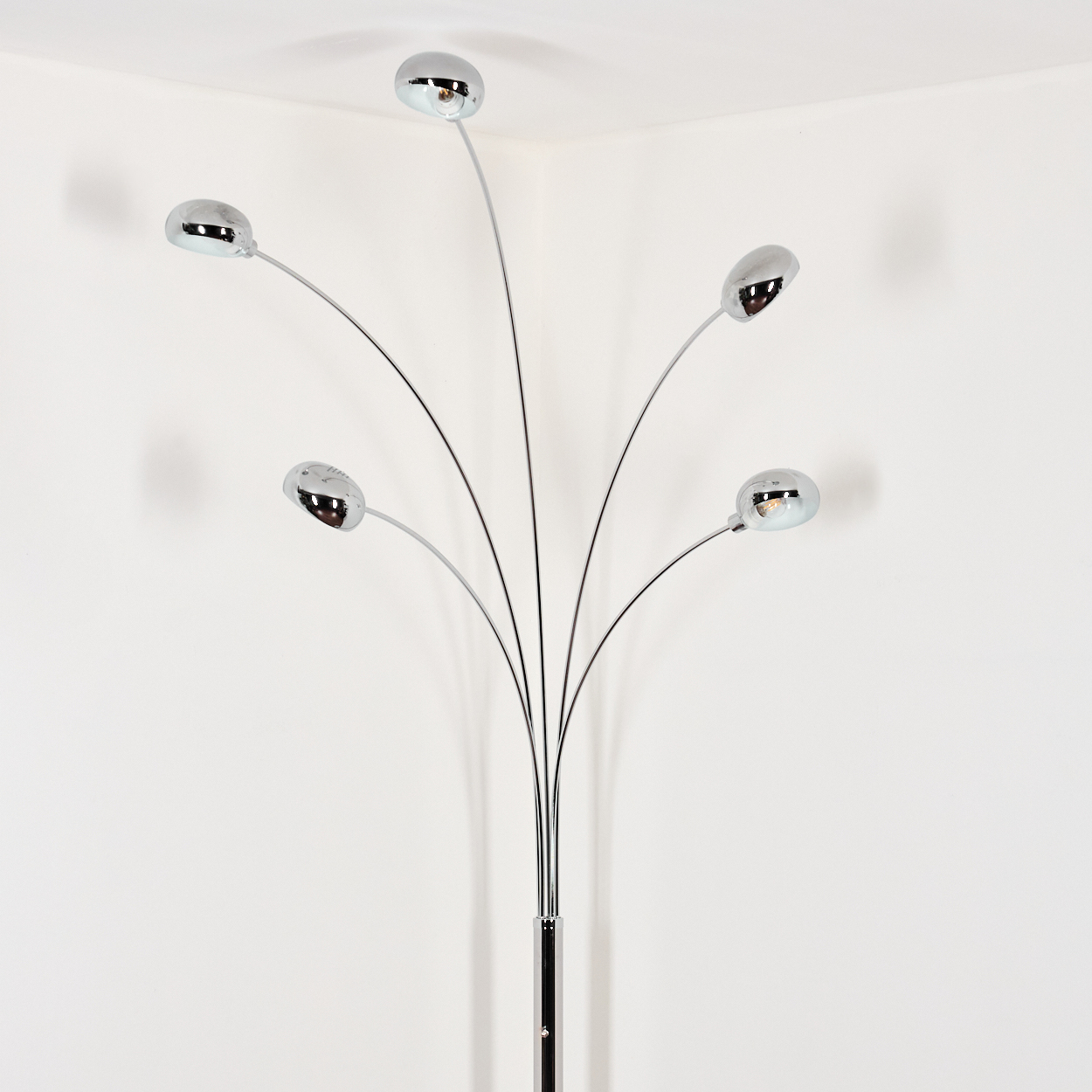 Eine verchromte Stehlampe mit fünf Lichtern. Der Sockel ist schwarz. Sie steht in der Ecke eines weißen Raumes und wirft Schatten.