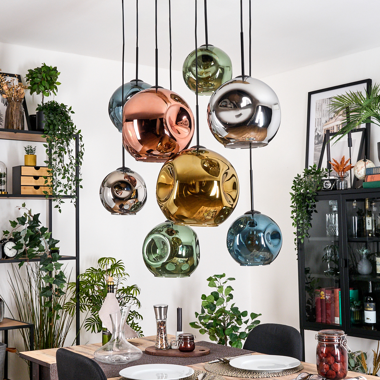 Esszimmer mit Holztisch, hängenden kugelförmigen Lampen in verschiedenen Farben und Pflanzen. Ein schwarzer Schrank und Bilderrahmen im Hintergrund.