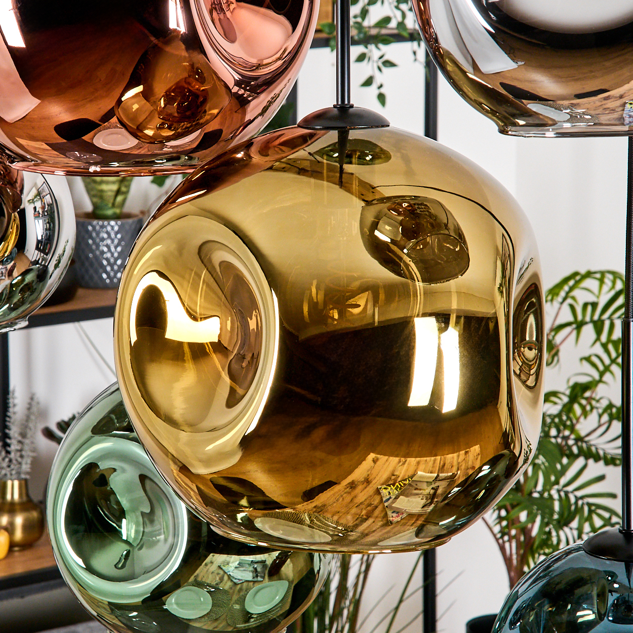 Nahaufnahme von modernen, kugelförmigen Pendelleuchten in Gold, Kupfer, Grün und Blau. Reflexionen zeigen die Umgebung mit Pflanzen im Hintergrund.