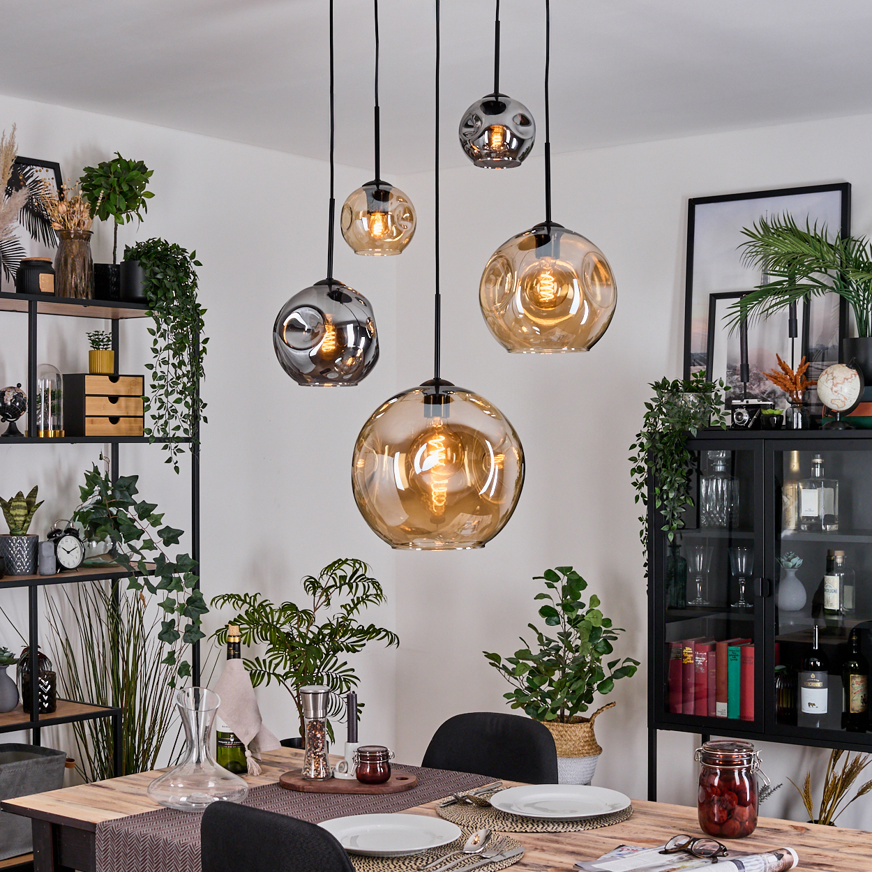 Esszimmer mit hängenden Kugelleuchten. Tisch für Mahlzeit gedeckt. Regale mit Pflanzen und Dekoration. Schwarzer Schrank. Pflanzen.