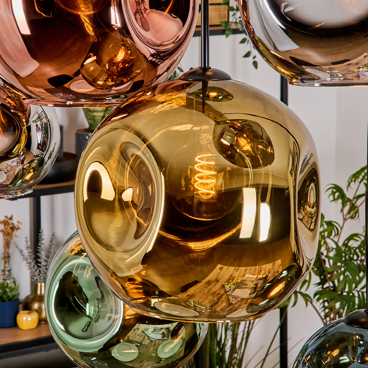 Nahaufnahme von modernen Pendelleuchten mit Glaskugeln in Gold, Kupfer und Grün. Innenraum mit Pflanzen und Regalen sichtbar.
