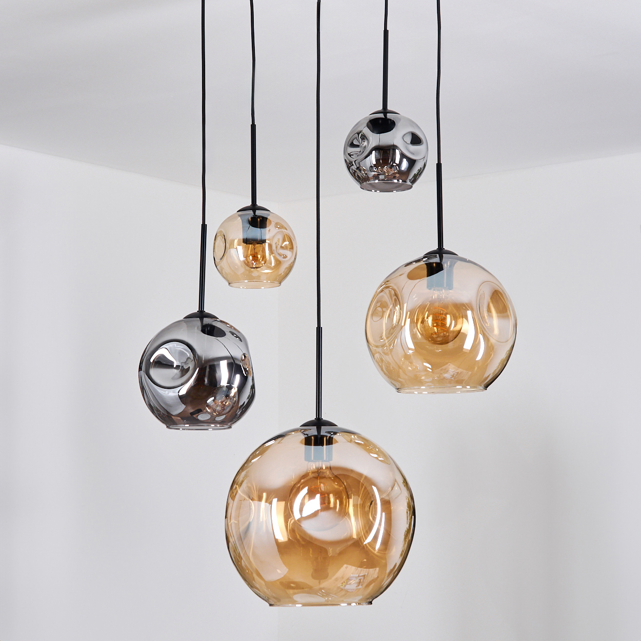 Fünf hängende Pendelleuchten. Einige sind aus bernsteinfarbenem Glas, andere aus Chrom. Schwarze Kabel hängen sie von einer weißen Decke ab, in einer Ecke.