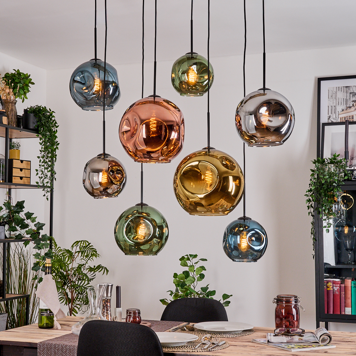 Mehrere bunte Glas-Pendelleuchten hängen über einem Holztisch, Pflanzen stehen auf Regalen im Hintergrund.