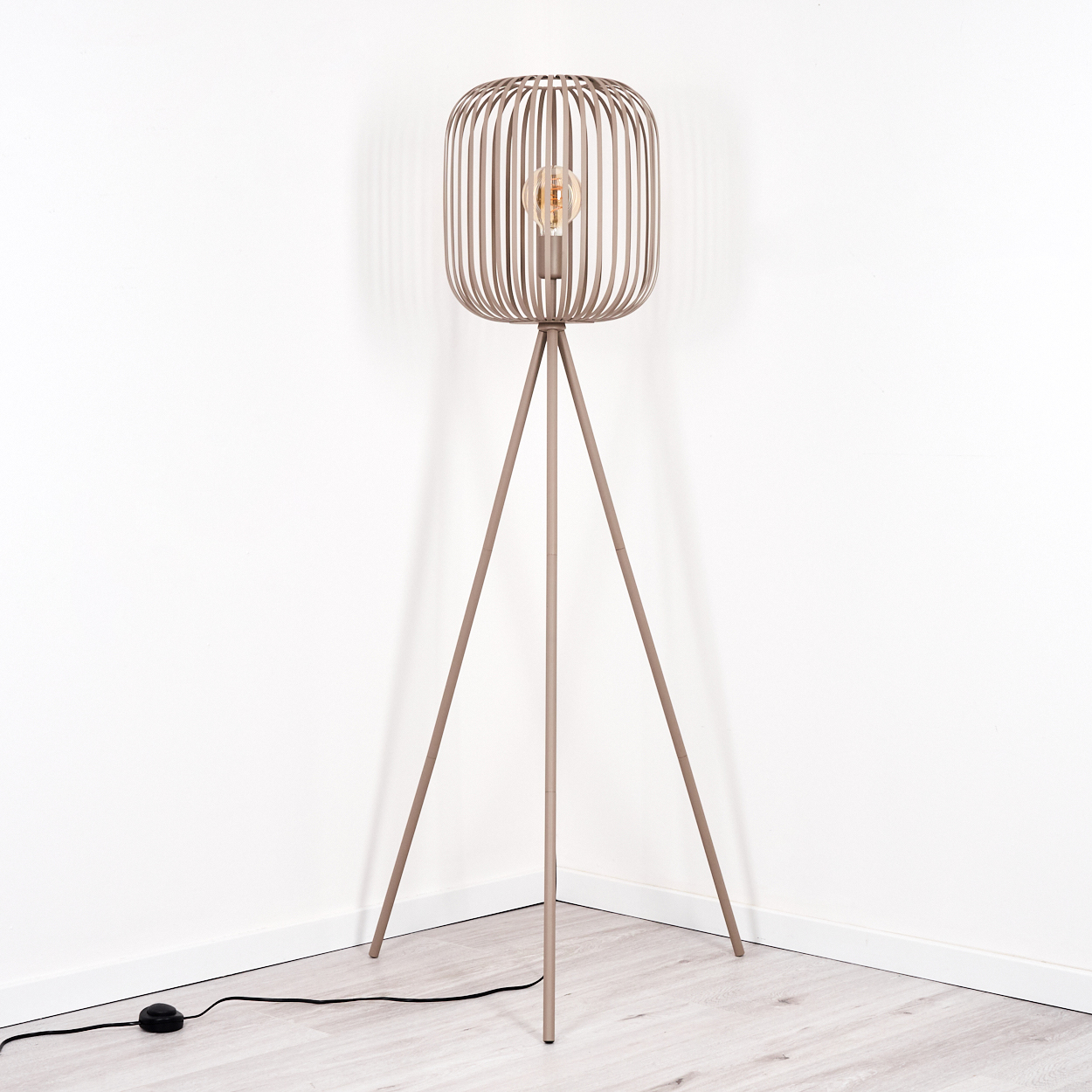 Eine moderne Stehlampe mit käfigartigem Lampenschirm, sichtbarer Glühbirne und Dreibein-Sockel steht in einer Ecke vor einer weißen Wand.