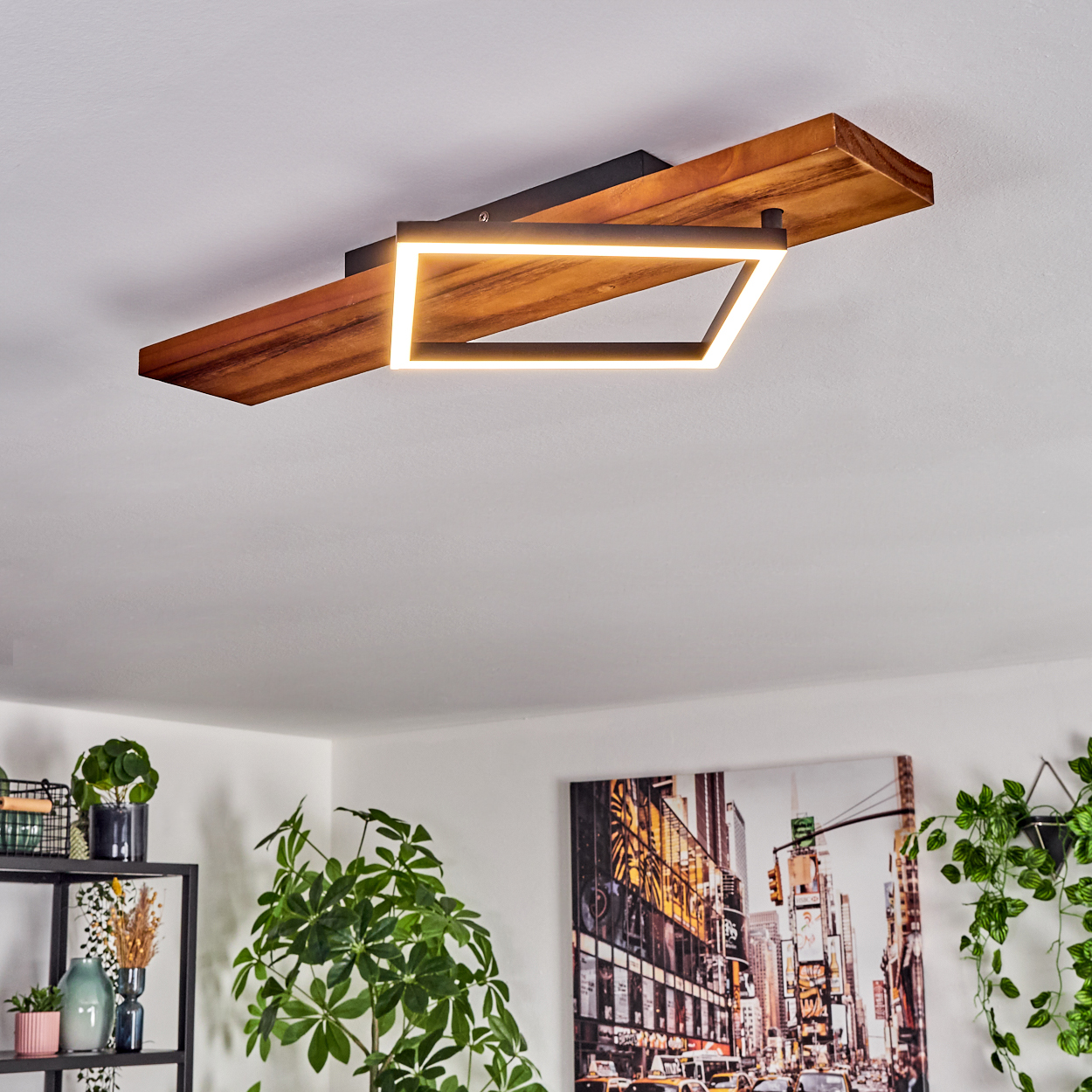 Eine moderne Deckenleuchte mit Holzsockel und quadratischer Leuchte. An der Wand hängt ein Stadtbild.