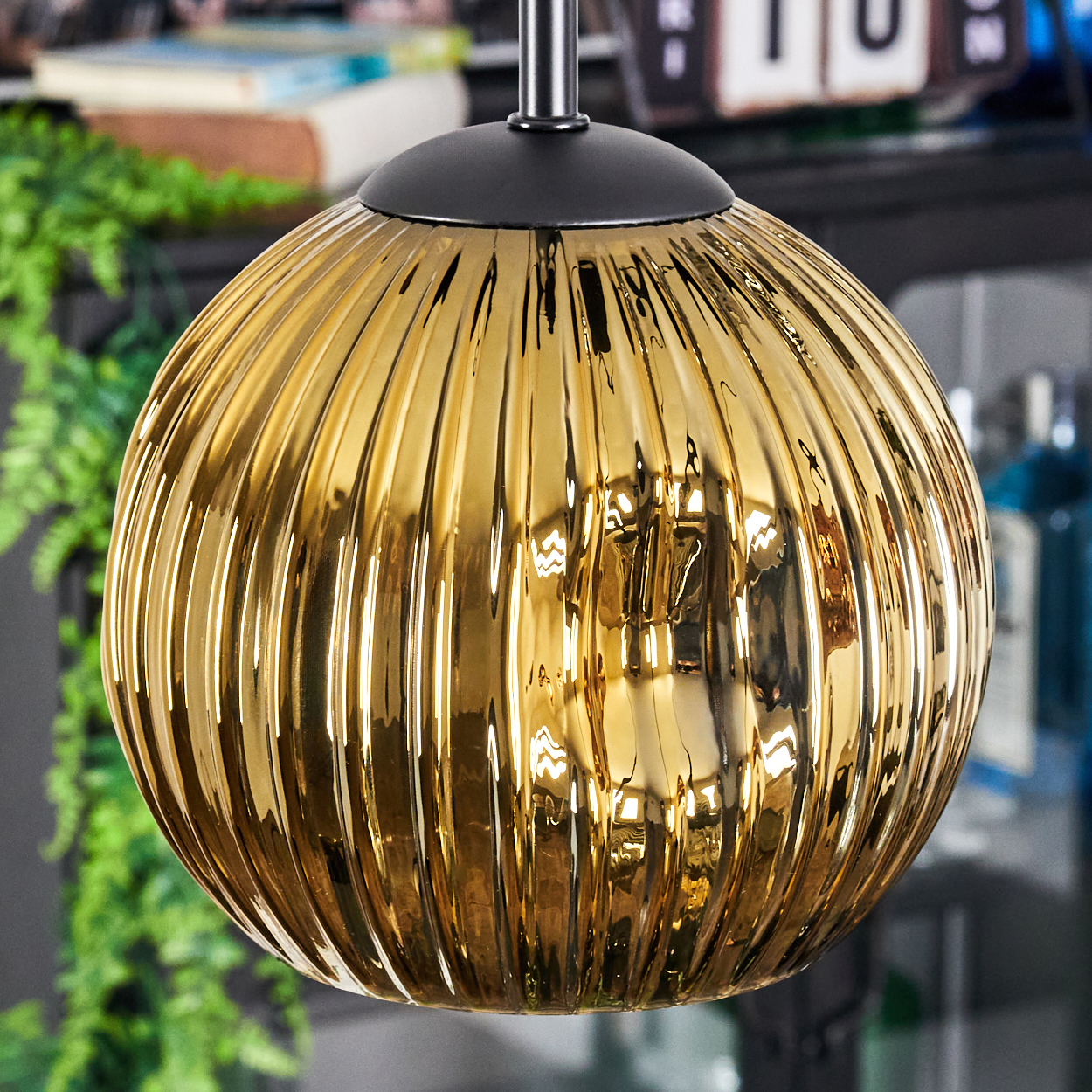 Eine goldene, gerippte Glas-Pendelleuchte hängt von der Decke. Im Hintergrund sind grüne Blätter und Bücher zu sehen.