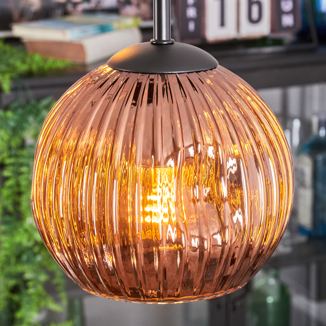 Eine kupferfarbene, gerippte Glas-Hängeleuchte ist beleuchtet. Der Hintergrund umfasst Grün, einen Kalender und Regale mit Flaschen.