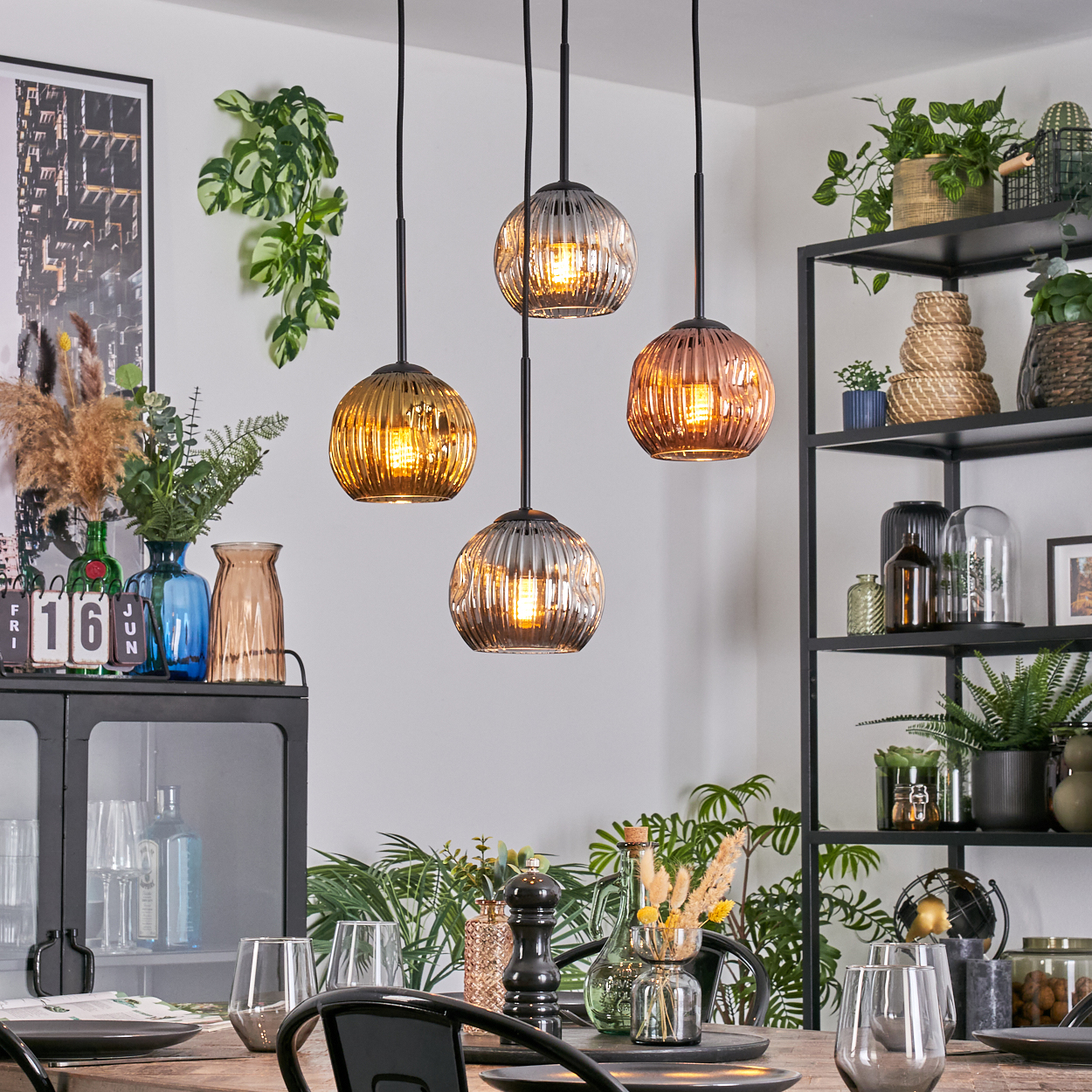 Esszimmer mit Hängelampen, Regalen mit Pflanzen, gedecktem Tisch und Dekorationsartikeln. Warme Töne und natürliches Licht.
