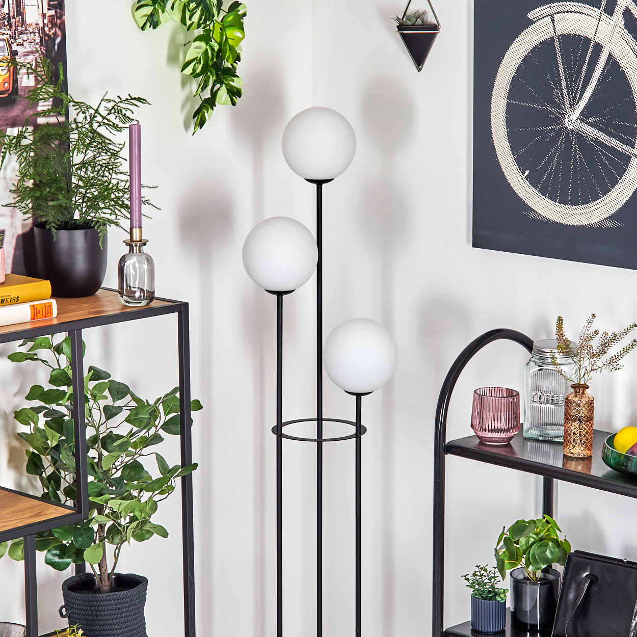 Eine Zimmerecke mit einer Stehlampe mit drei weißen Kugeln. Regale zeigen Pflanzen, Bücher und dekorative Gegenstände. Fahrradkunst an der Wand.
