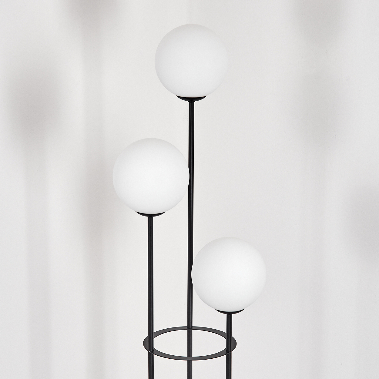 Eine moderne Stehlampe mit drei kugelförmigen weißen Glühbirnen an schwarzen Stielen. Der Hintergrund ist eine weiße Wand mit Schatten.